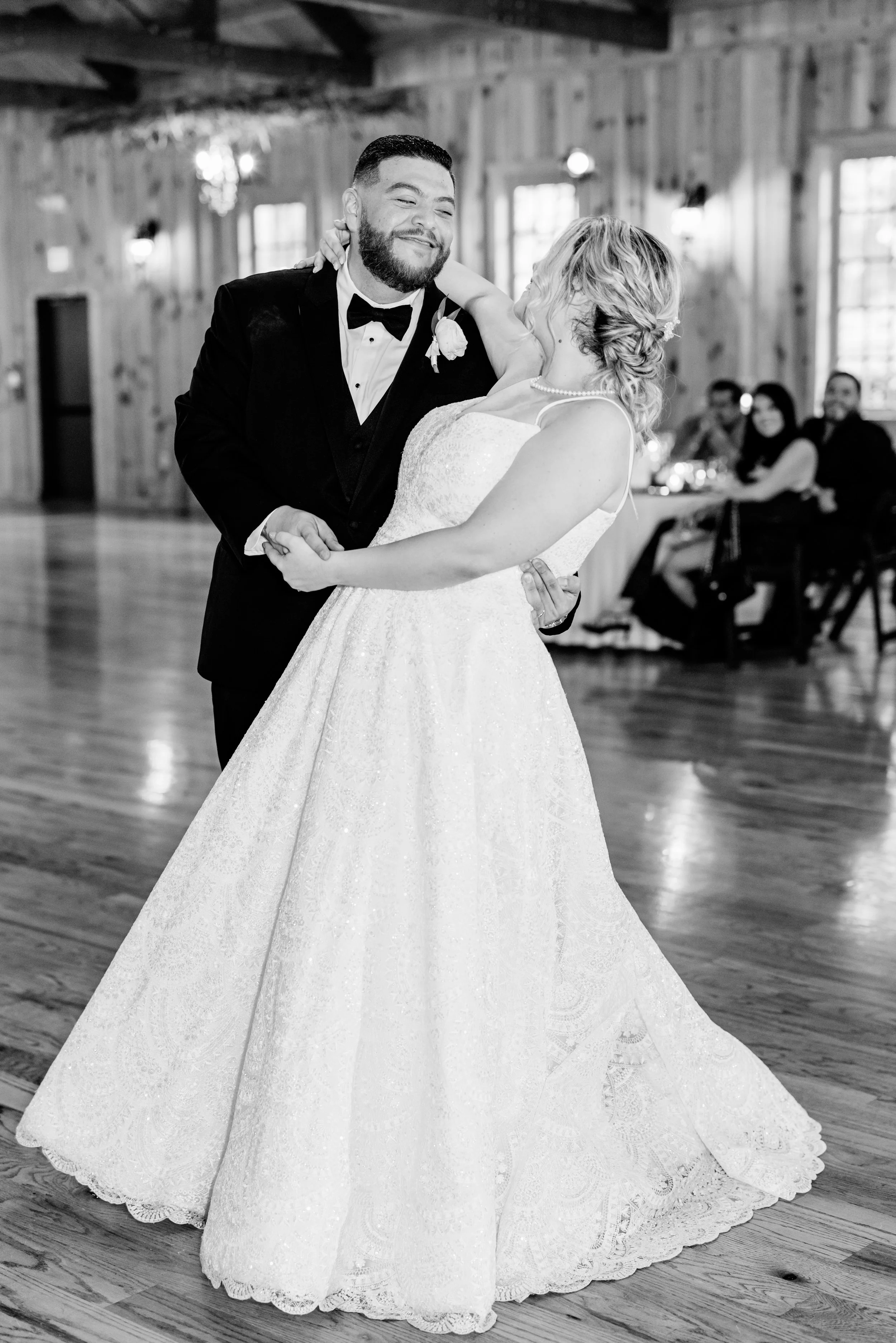 A bride and groom dancing at their wedding reception. The bride is wearing a white gown and pearls, the groom is in a black tuxedo with a bow tie. They are smiling and looking at each other. The background shows other guests seated at tables in a rustic wooden venue.