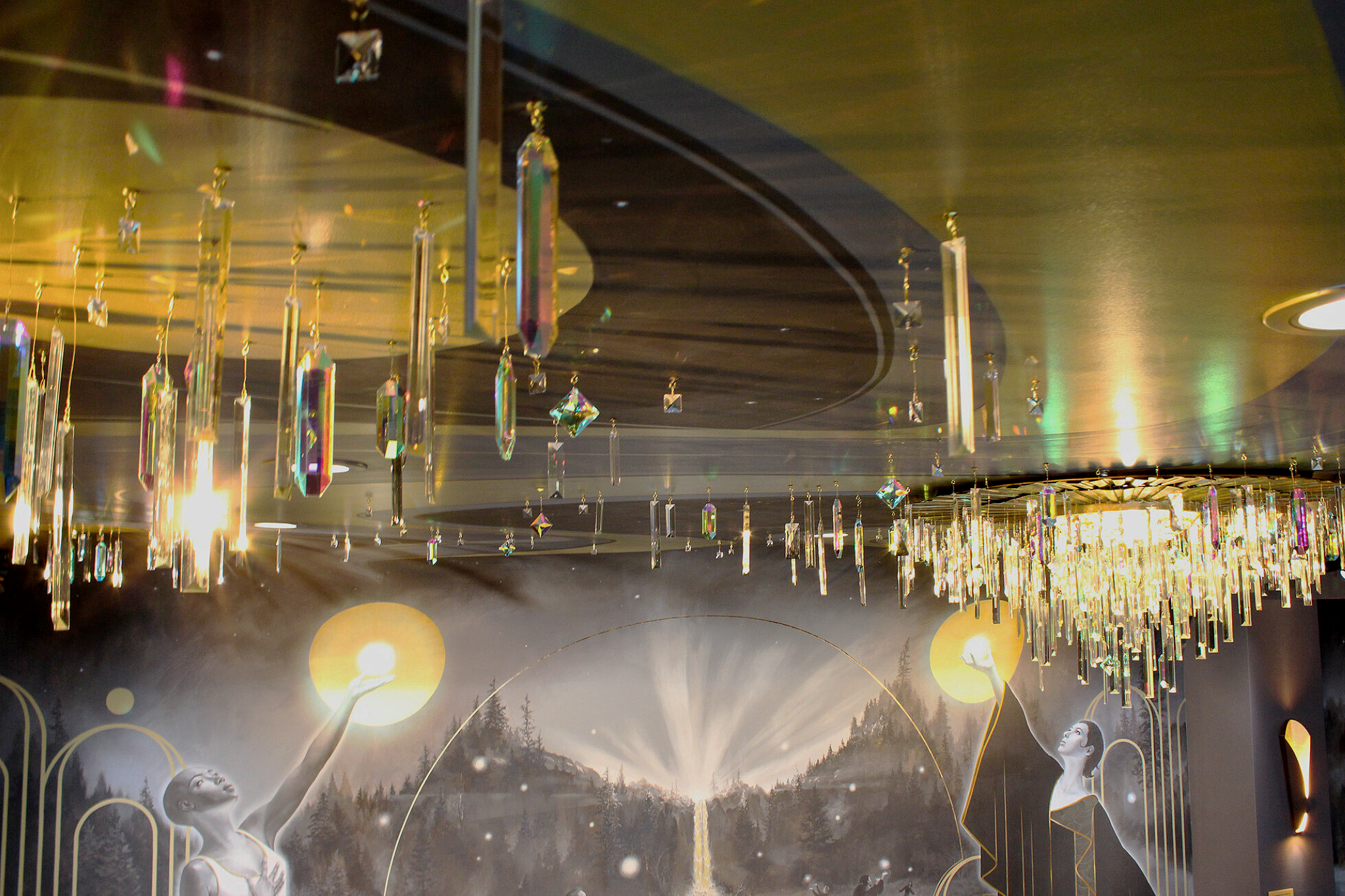 Interior of an elegant room with a large, ornate chandelier decorated with hanging crystal prisms, and a mural featuring two women reaching towards glowing yellow orbs against a mountainous landscape background.