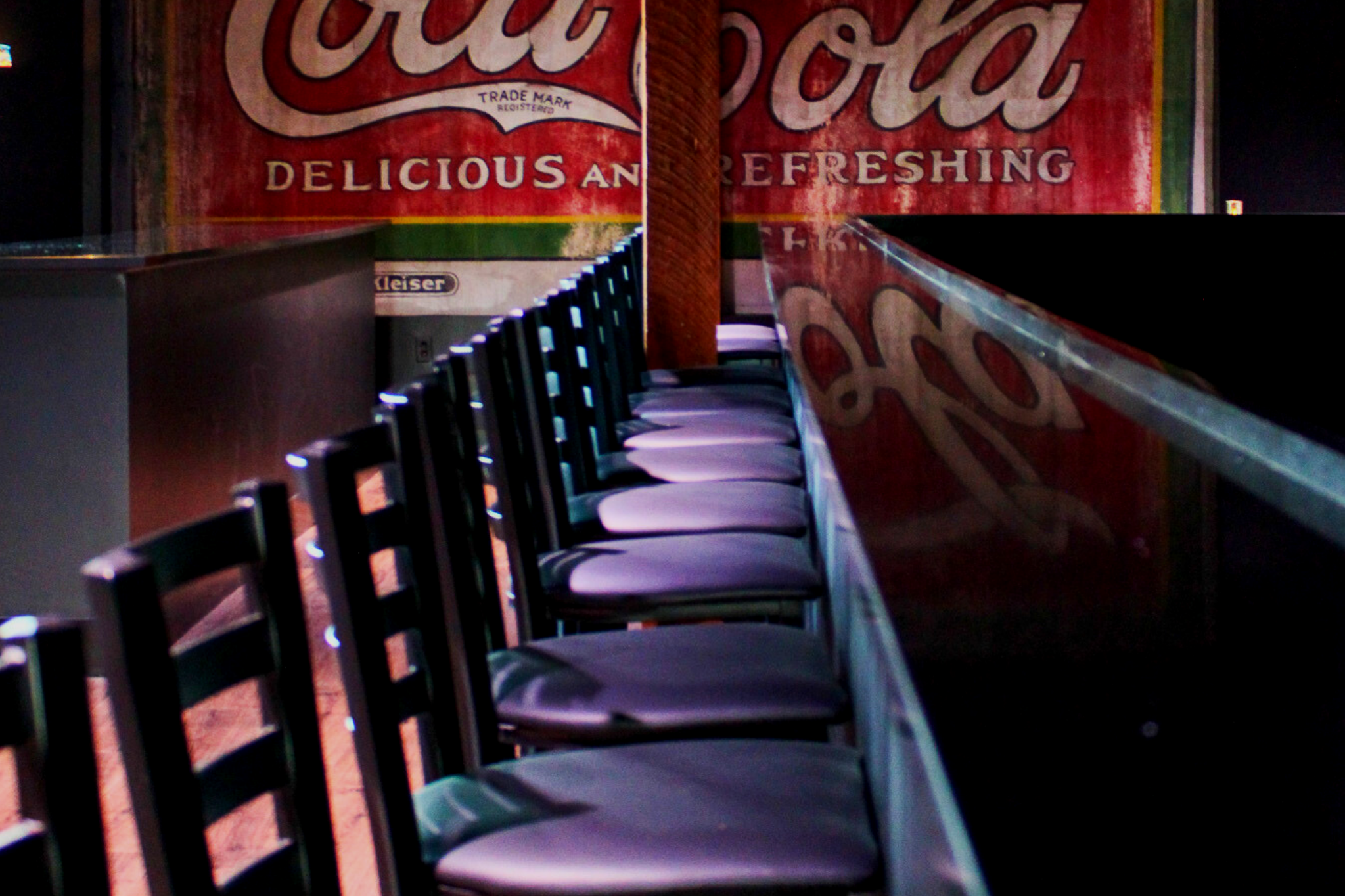 Empty bar stools lined up in front of a bar counter with a vintage Coca-Cola advertisement on the wall in the background.