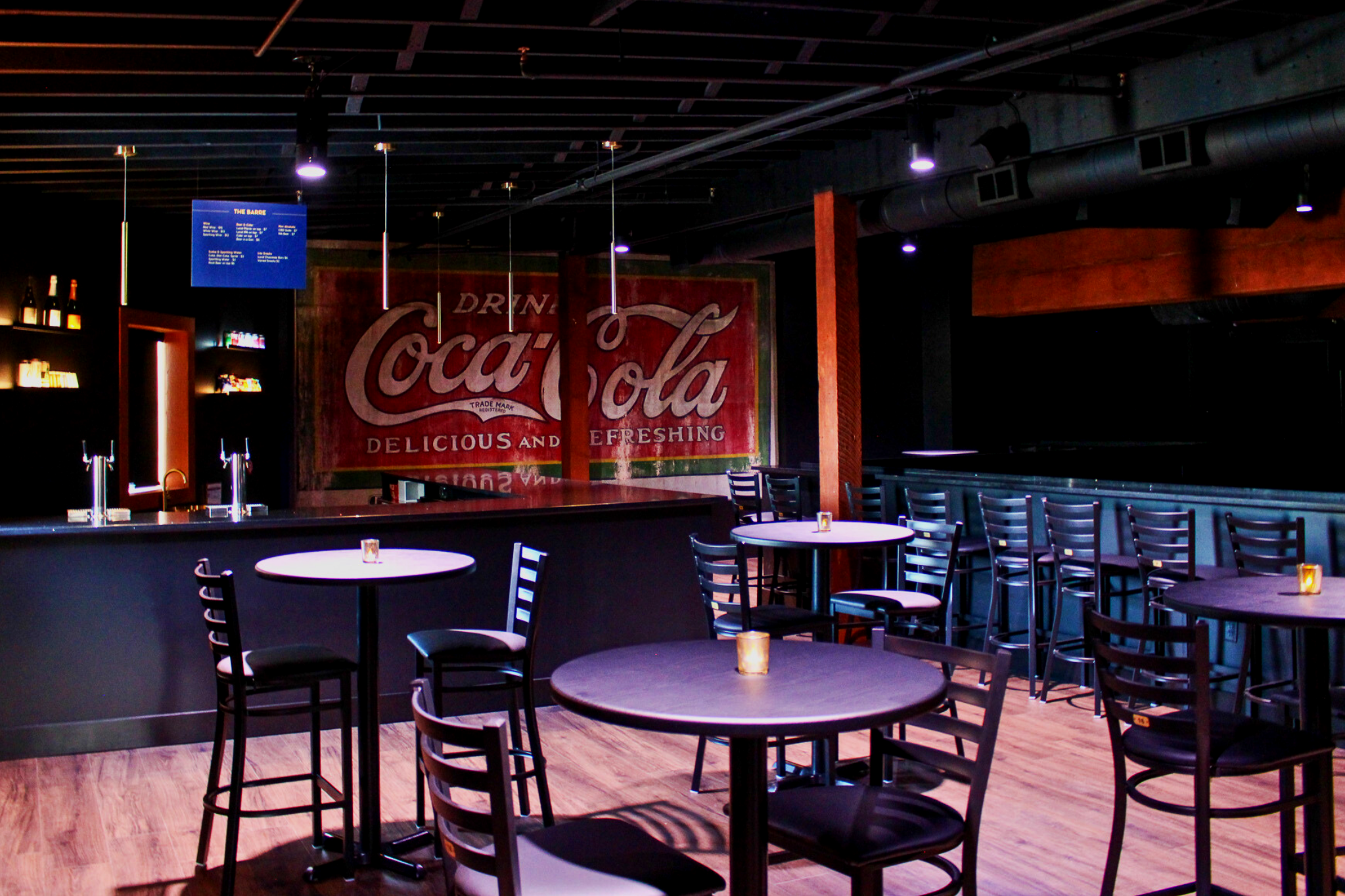 Empty bar with a Coca-Cola sign on the wall, round tables with chairs, and dim purple lighting.