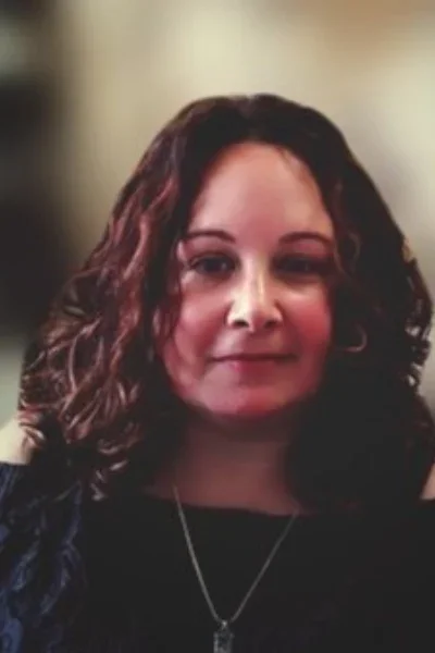 A woman with curly dark brown hair, wearing a black top and a necklace, looking at the camera with a slight smile.