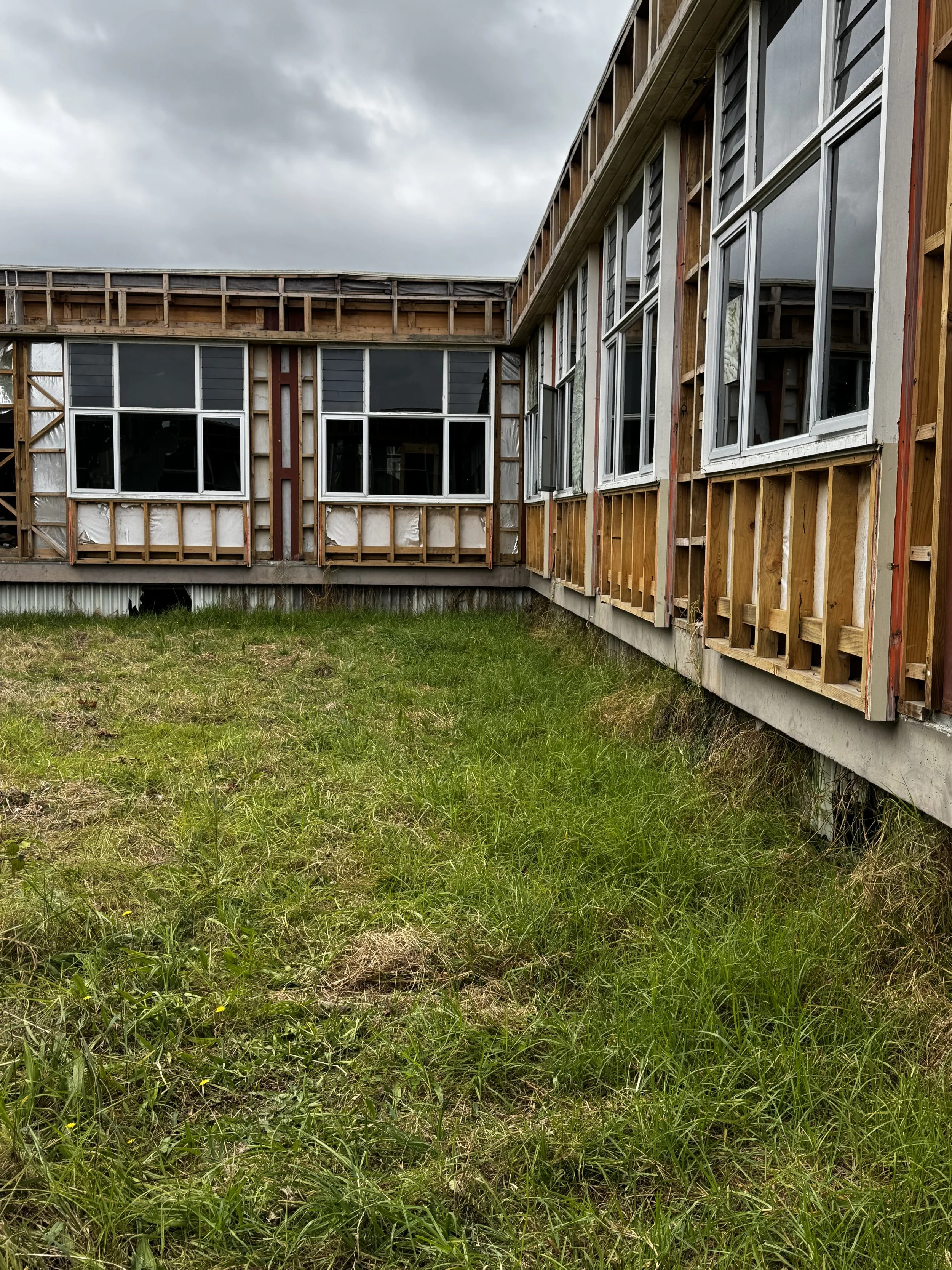 An unfinished house with exposed wooden framing and large windows, with a grassy lawn in the foreground and an overcast sky overhead.