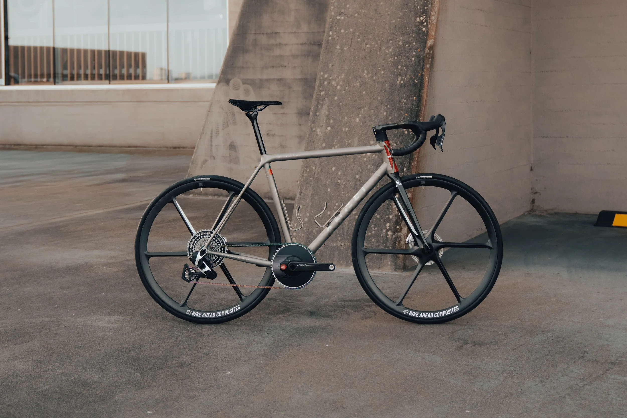 Bicicleta de carreras con marco gris y ruedas de fibra de carbono, estacionada frente a una pared de concreto en un estacionamiento.