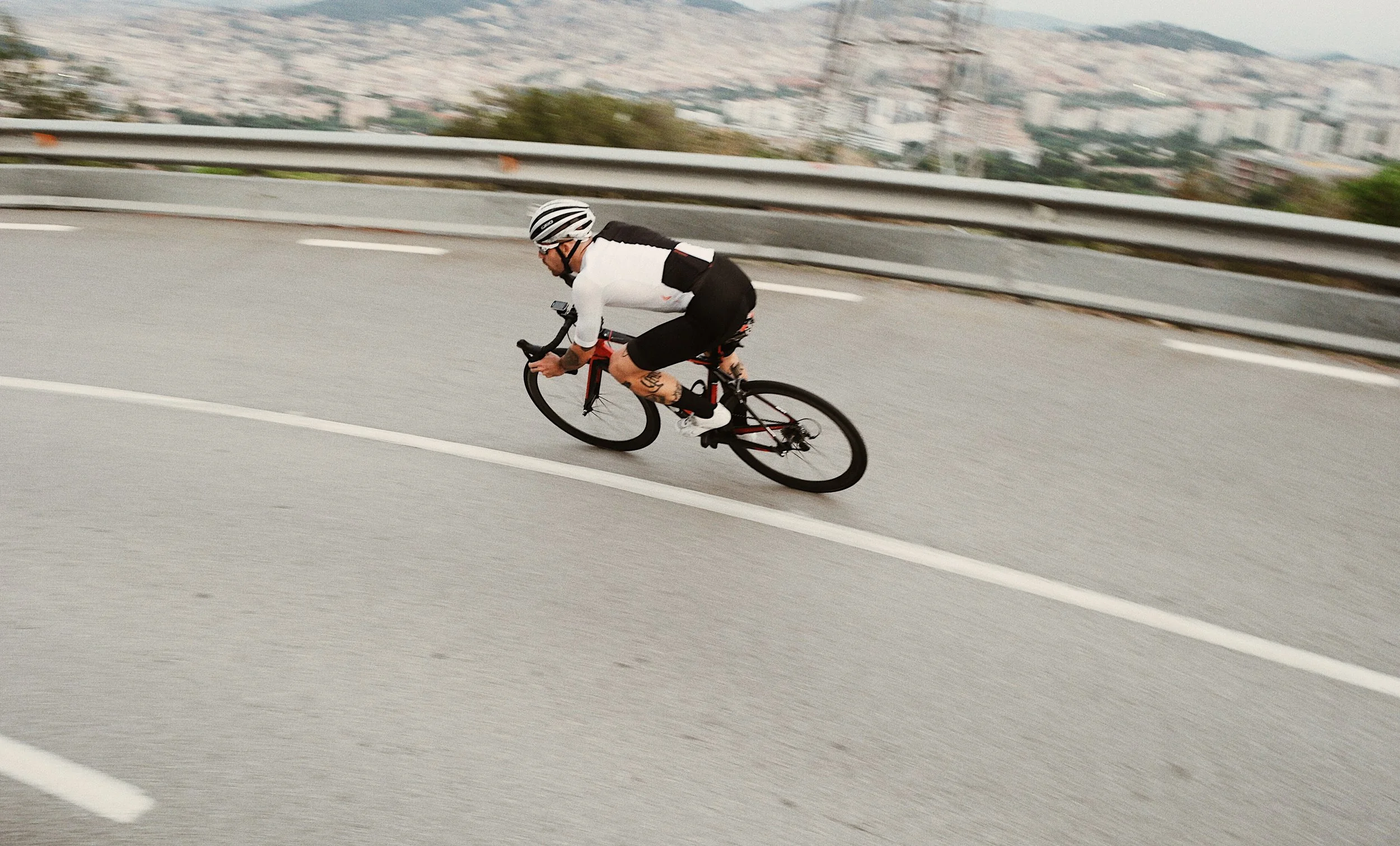 Un ciclista vestido con ropa blanca y pantalones negros, usando casco, pedaleando en una curva de una autopista elevada con vista a la ciudad.