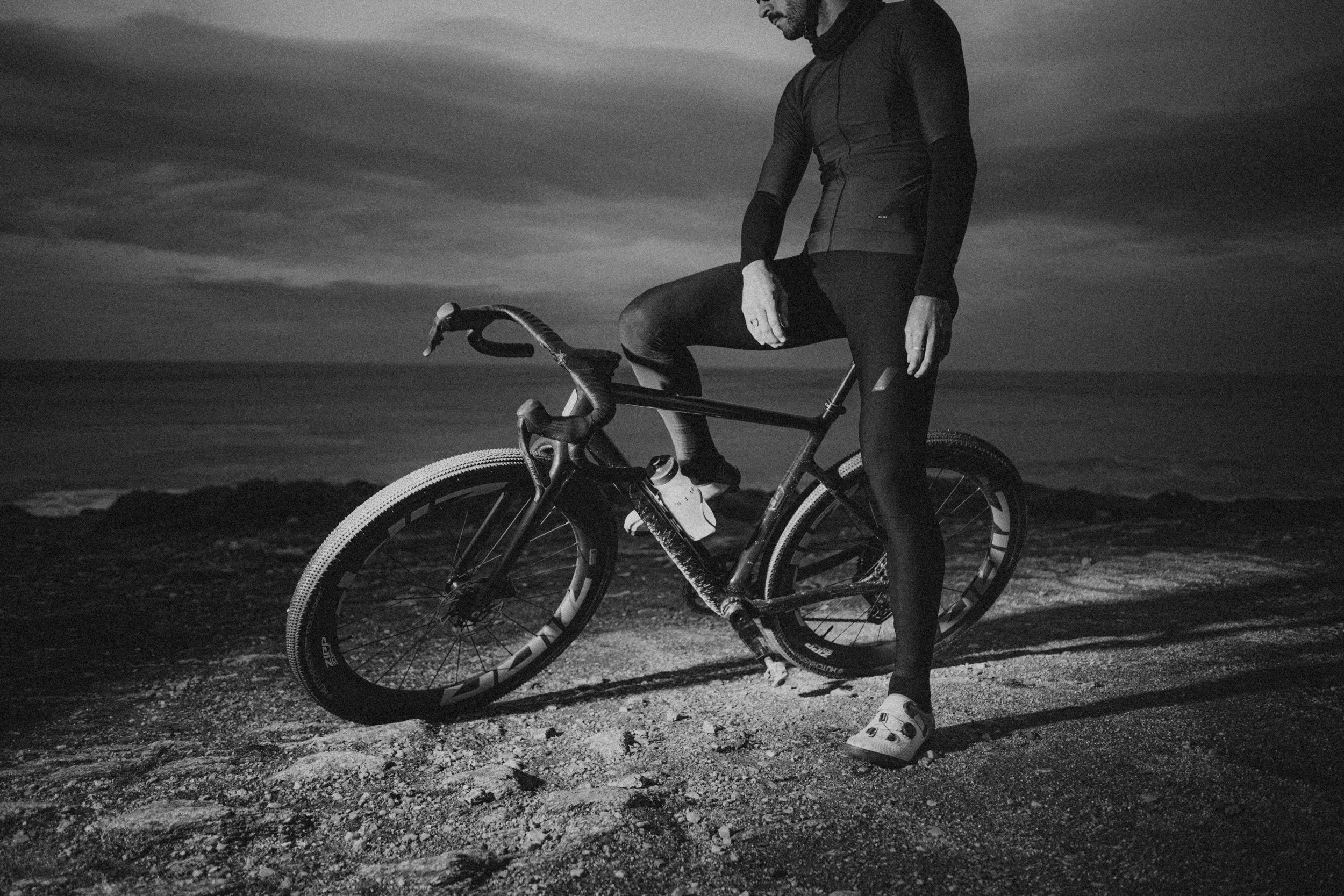 Persona con ropa deportiva de ciclismo y casco de ciclismo, de pie junto a una bicicleta en un paisaje nocturno o al amanecer, en una zona arenosa o costera.