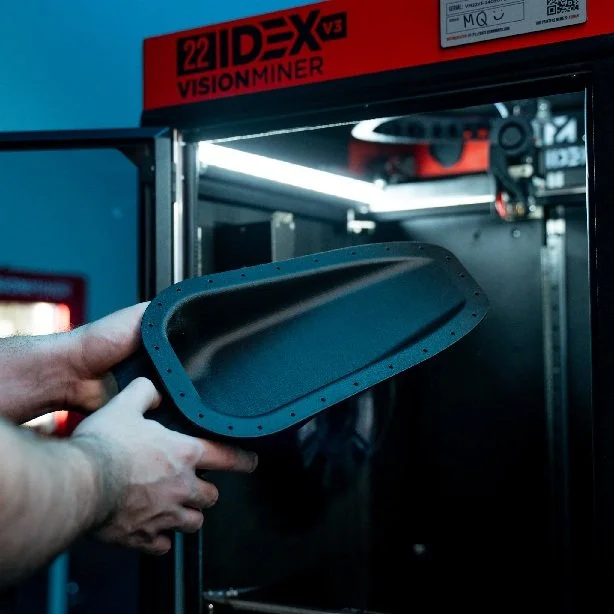 A person removes a large black 3D-printed part from a 22 IDEX V3 Vision Miner 3D printer inside a workshop setting, showcasing industrial additive manufacturing capabilities.