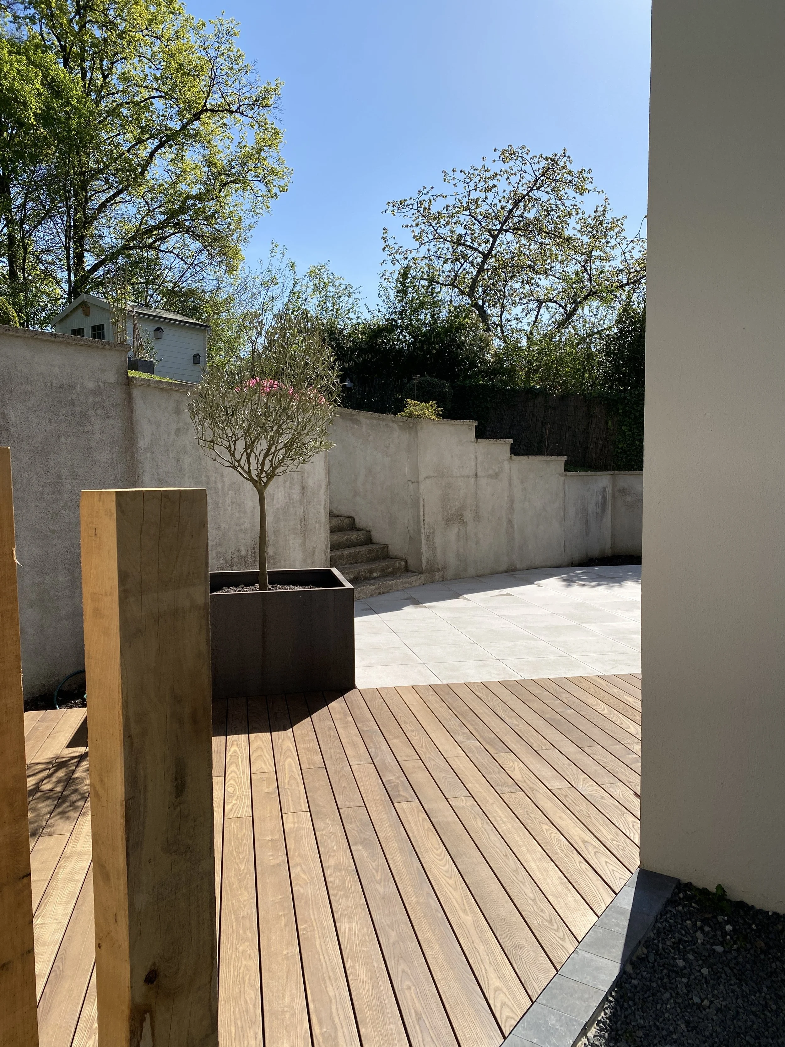 Cour ensoleillée avec terrasse en bois, pot de fleurs, escaliers en béton et mur blanc, entourée d'arbres et de végétation.