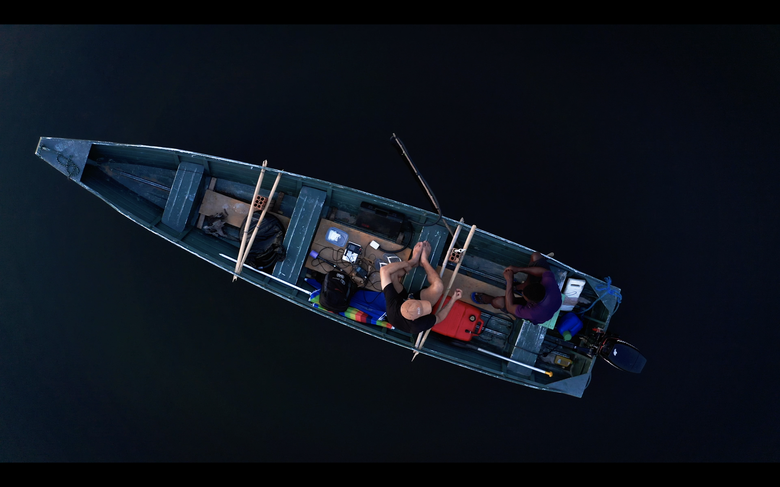 Overhead view of two people in a small wooden boat on dark water, with various equipment and oars on board.