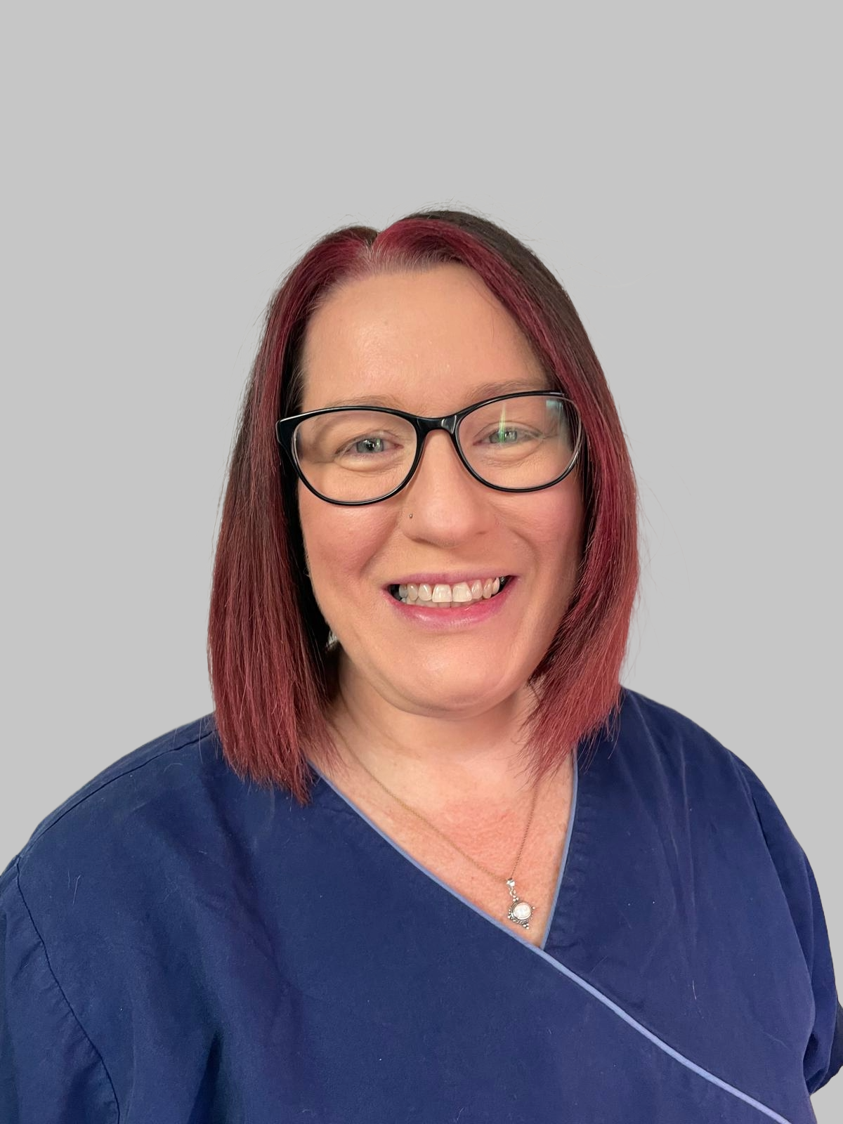 A woman with shoulder-length burgundy hair, wearing glasses, a blue medical scrub top, and a silver necklace with a small pendant, smiling against a plain light grey background.