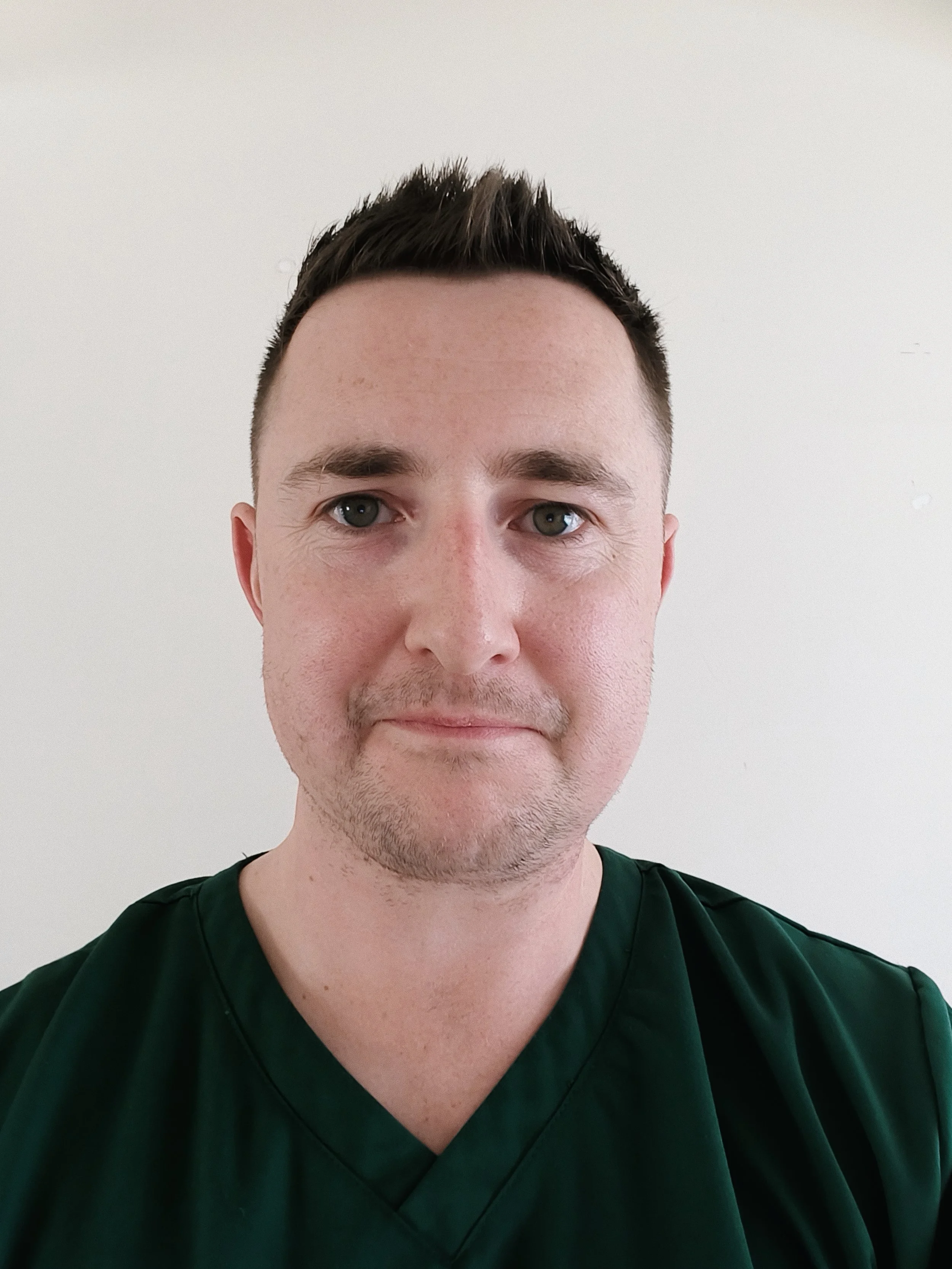 A young man with short brown hair, blue eyes, and light skin taking a selfie against a plain white wall. He is wearing a dark green shirt.