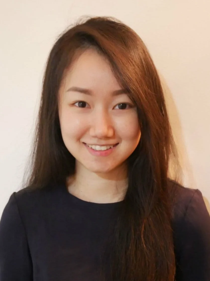 A young woman with long, dark hair and a smile, wearing a black top, standing against a plain light-colored background.