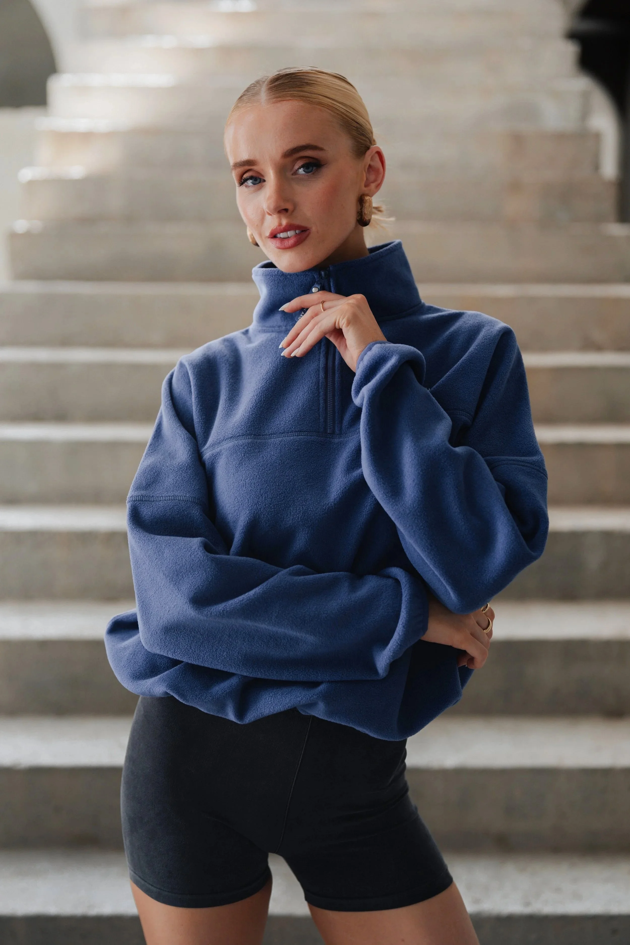 A woman with blonde hair in a ponytail, wearing a blue fleece quarter-zip top and black shorts, wearing 4th Arq Clothing.