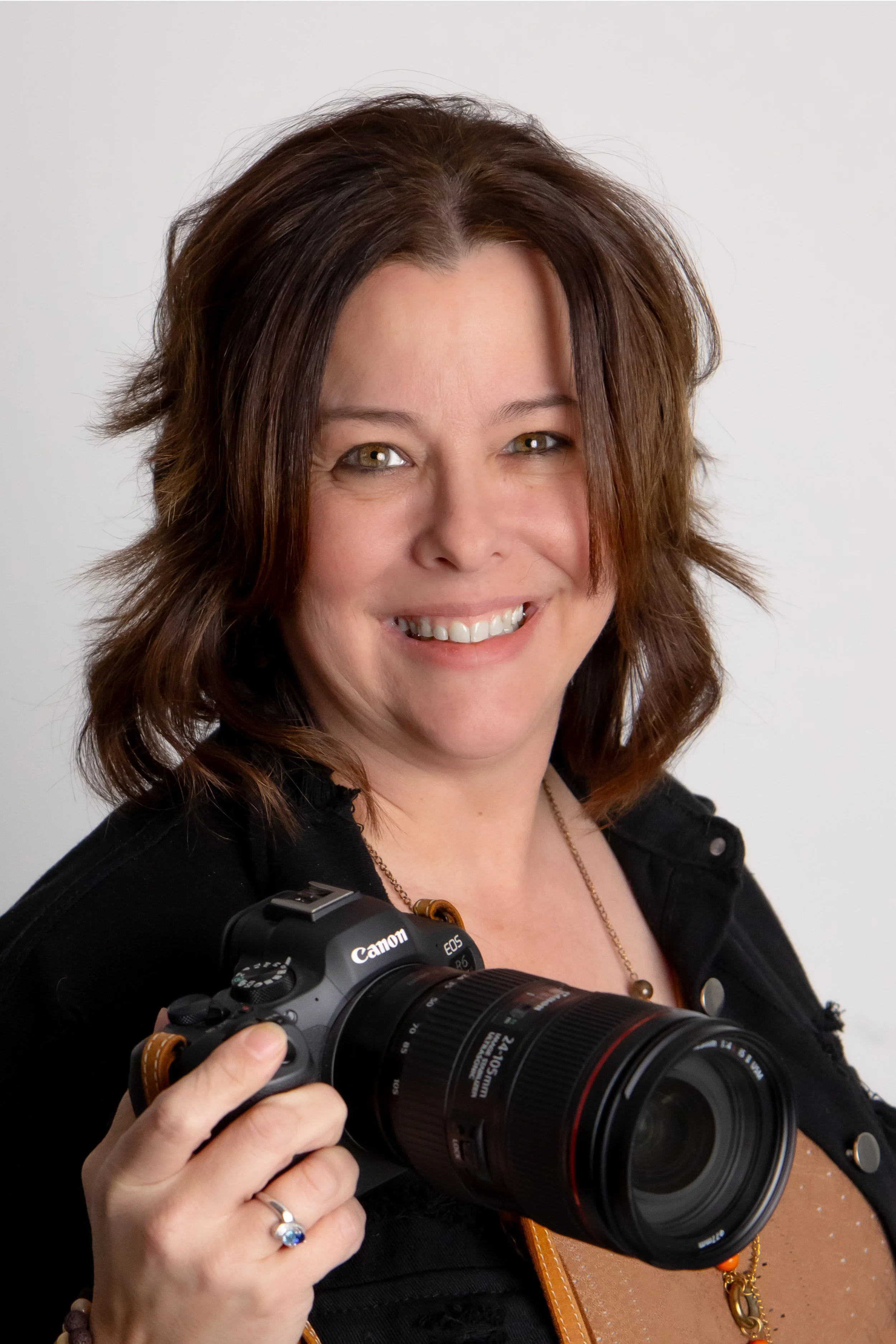 A woman with brown hair smiling and holding a Canon camera with a large zoom lens, wearing a black top, a ring on her finger, a necklace, and a lanyard, standing against a plain white background.