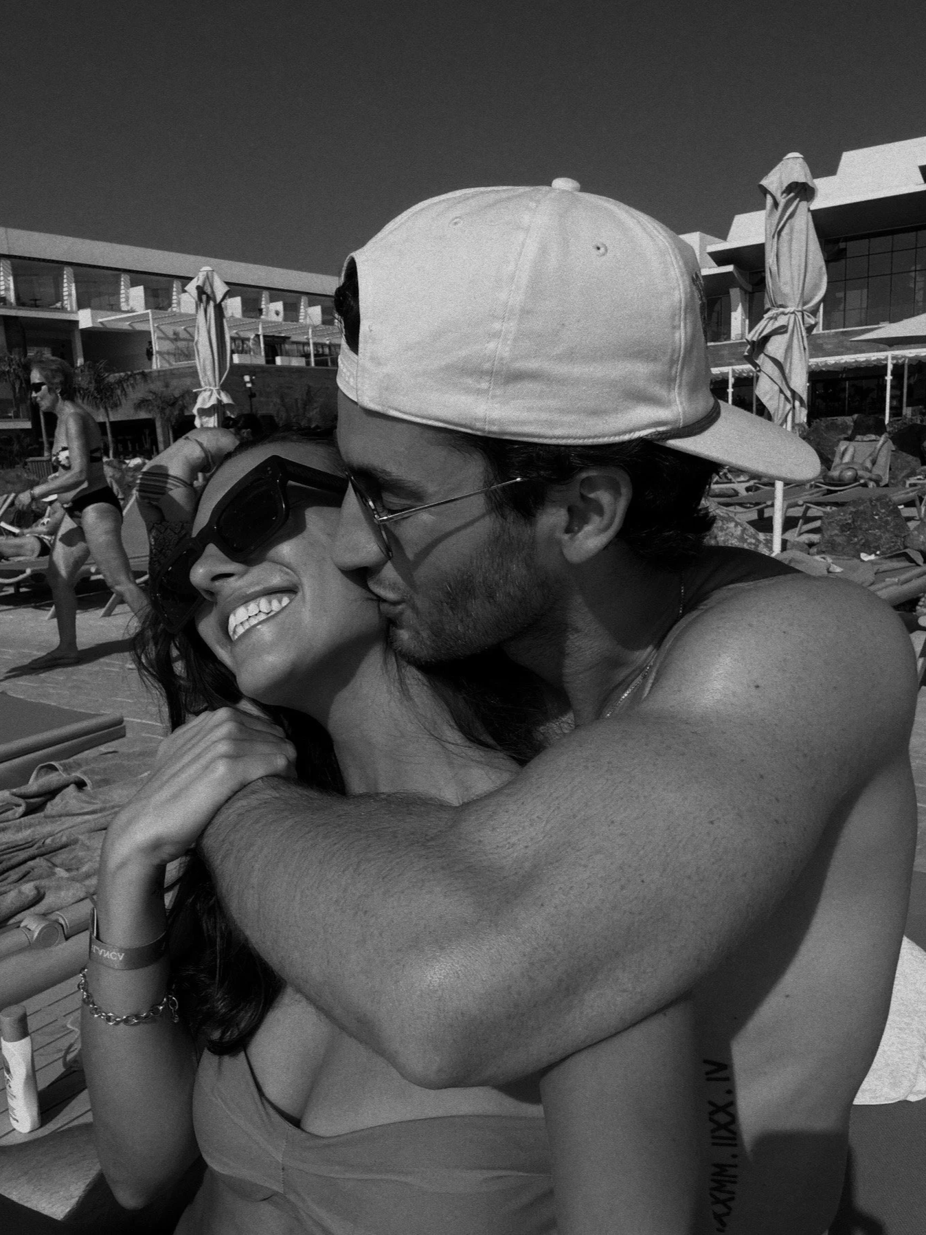 A smiling woman and a man, both wearing swimming attire, sitting close together on a beach chair, with the man kissing the woman's head. Background shows other beachgoers, umbrellas, and a modern building.