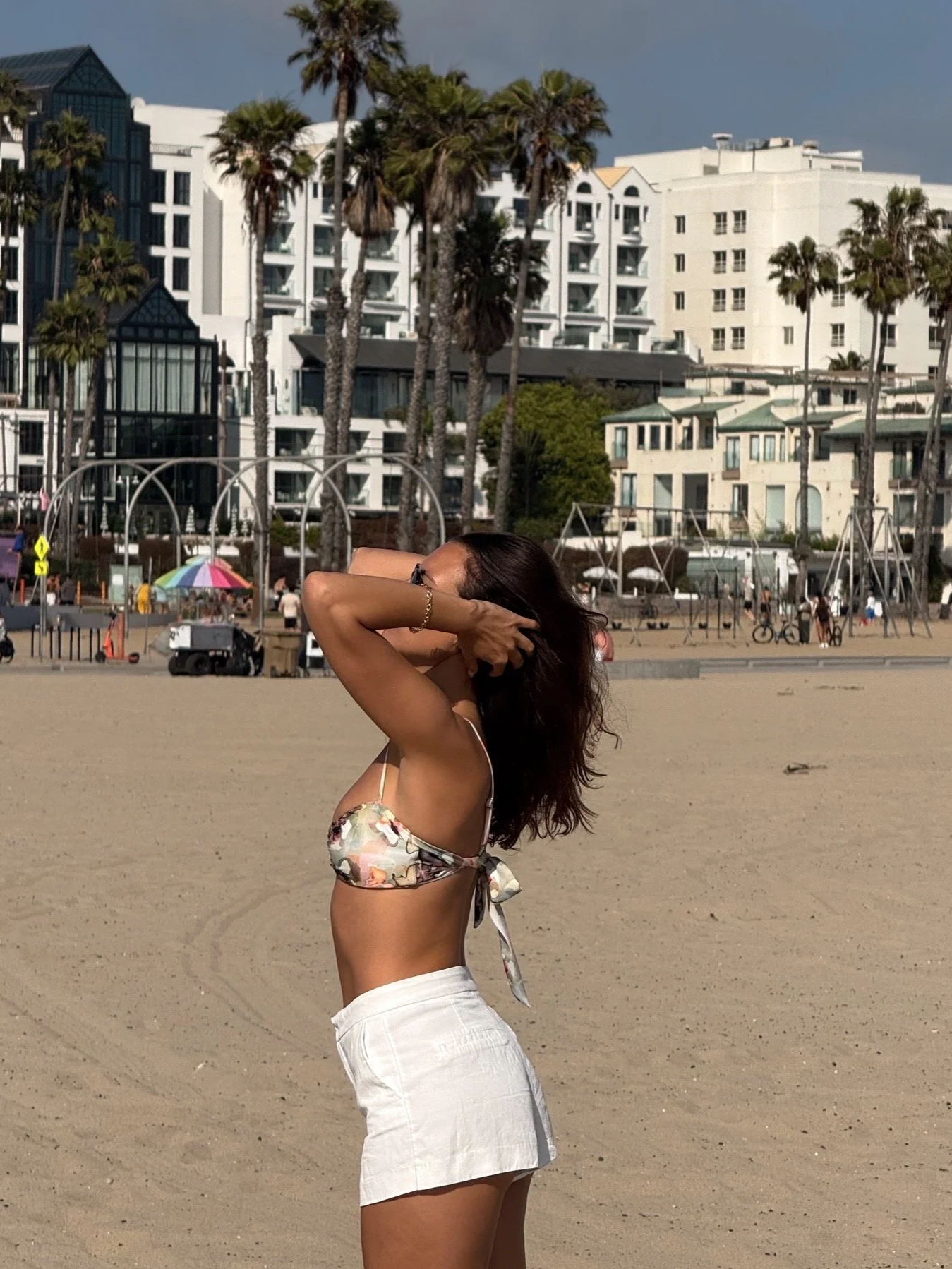 A woman in a floral bikini top and white shorts standing on a sandy beach with her hands behind her head. In the background, there are palm trees, playground structures, and multi-story buildings.
