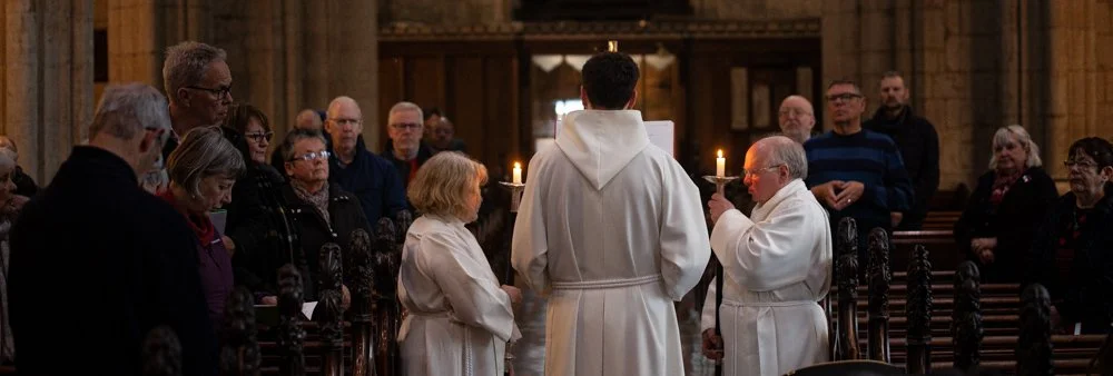 A server in a white robe holding a candle facing to the right. A vicar in a white robe with his back to the camera, and another server in a white robe facing to the left holding a candle to the