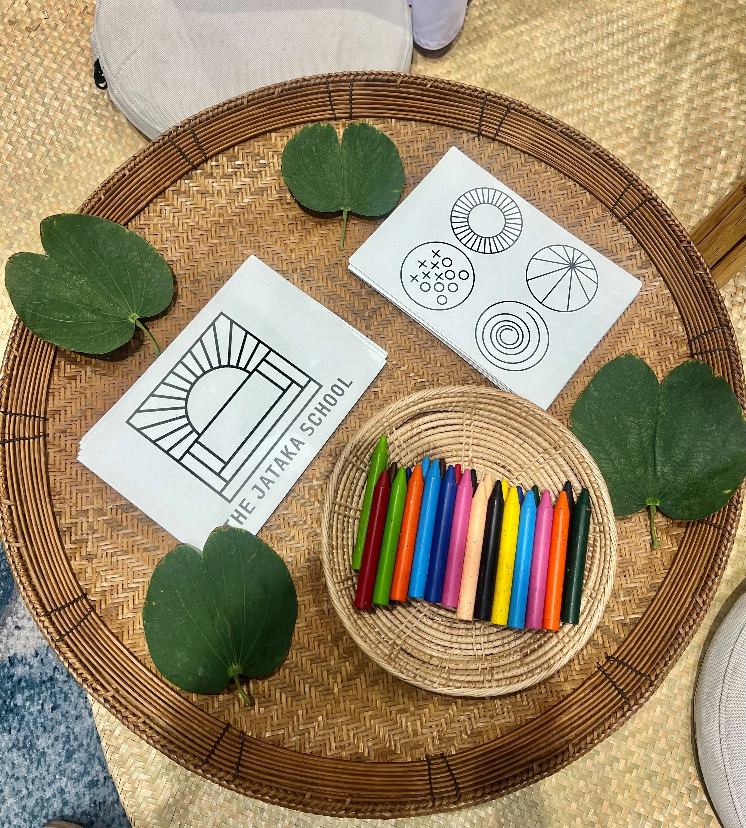A woven basket on a table with several green leaves and markers pens