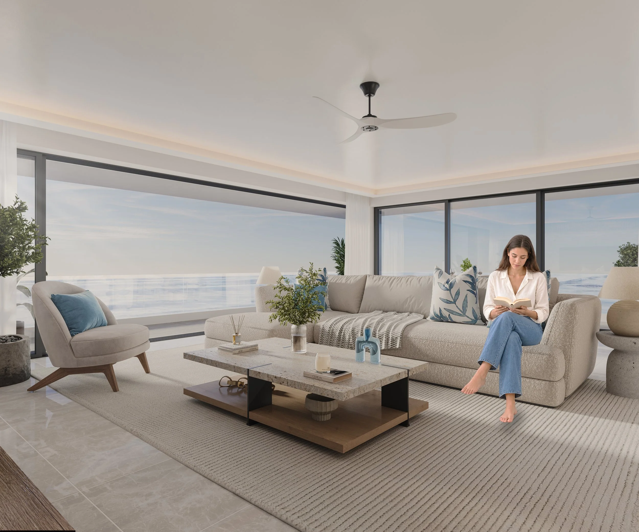 Modern living room with large floor-to-ceiling windows showing an ocean view, beige sofa with decorative pillows, a round armchair, a coffee table with decorative items and plants, and a ceiling fan.