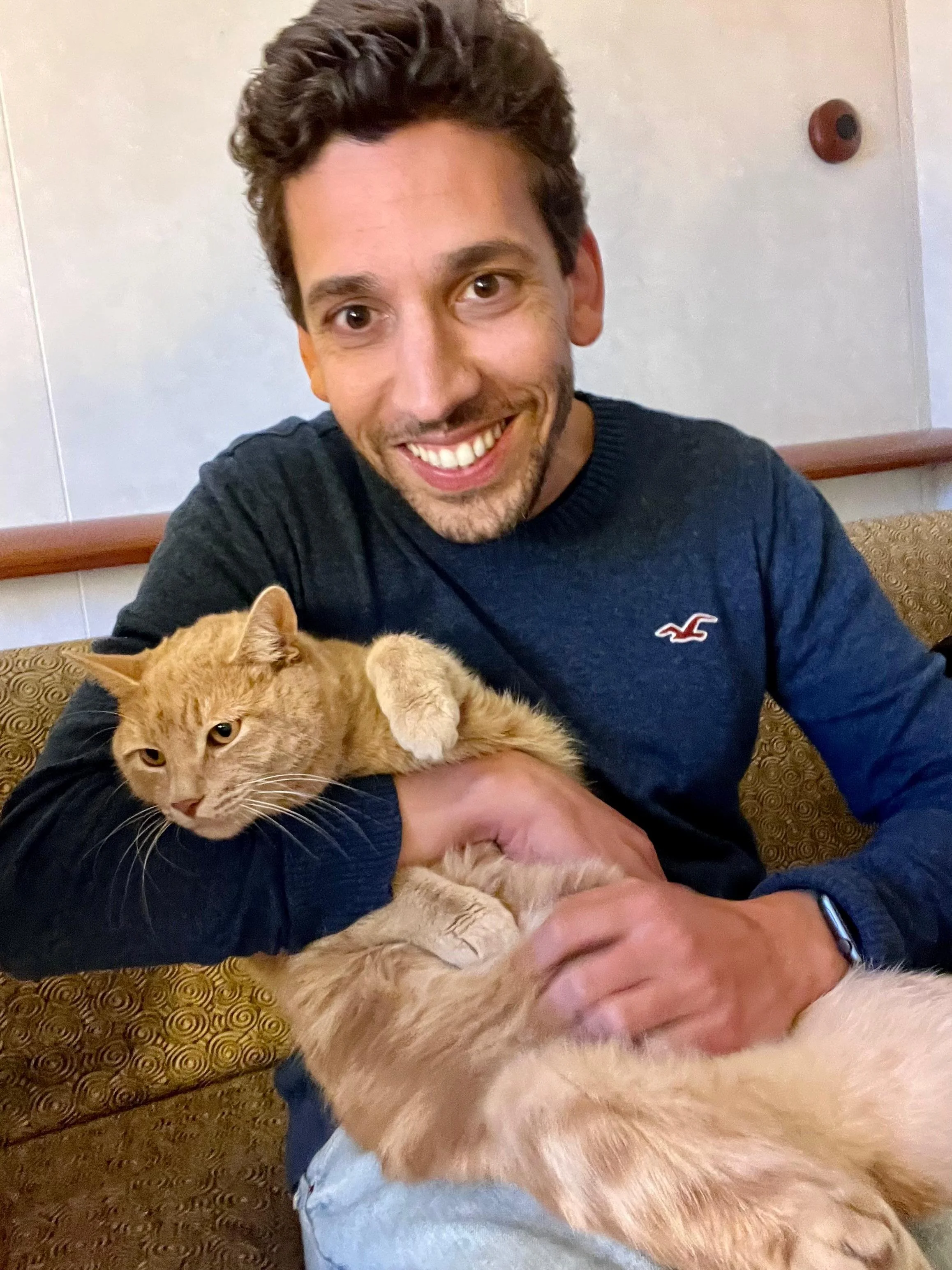A person smiling while holding a ginger cat on a couch.