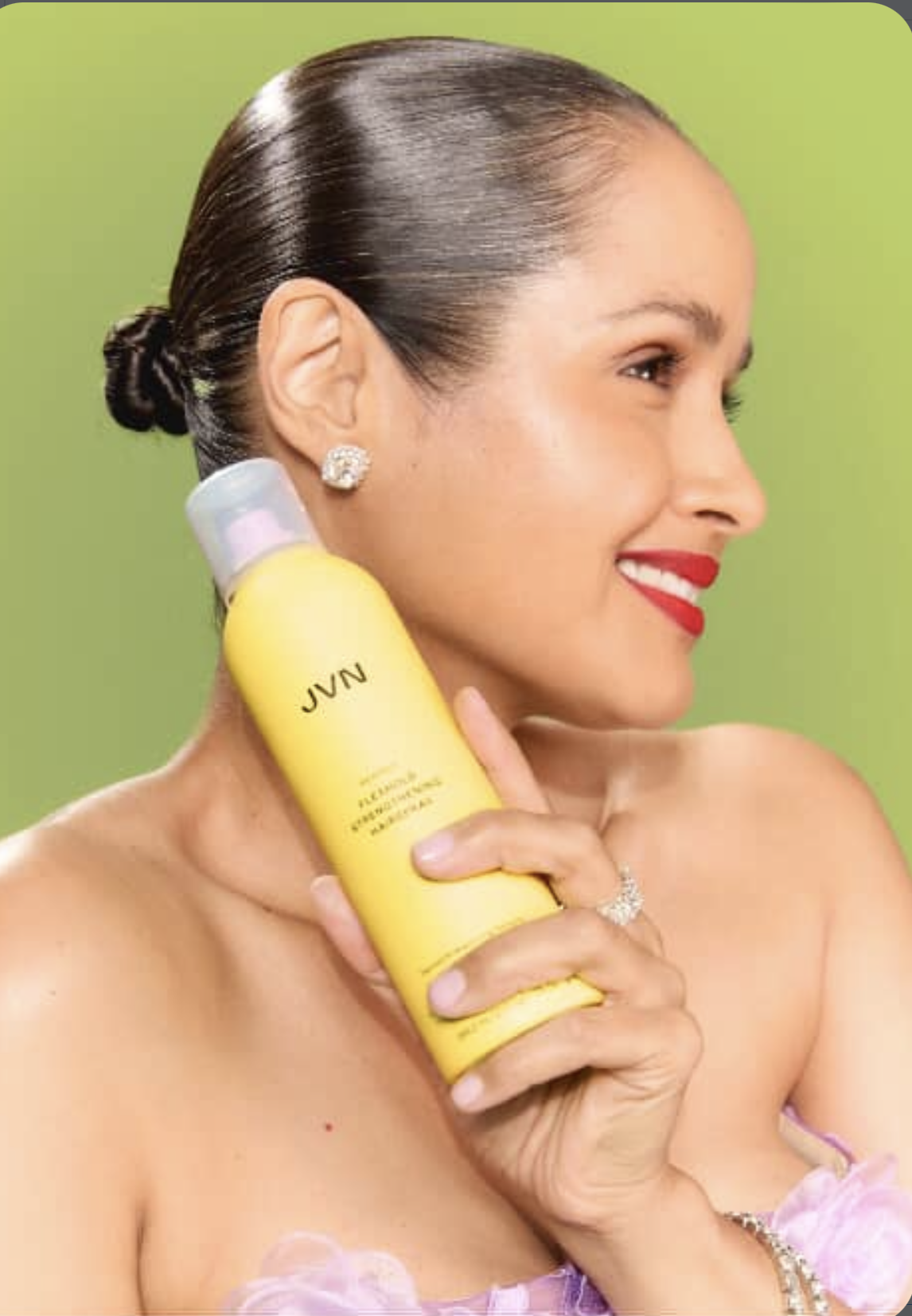 A woman with dark hair tied in a snatched baby hair bun, wearing red lipstick and diamond earrings, holding a yellow bottle of JVN hair hairspray against a green background.