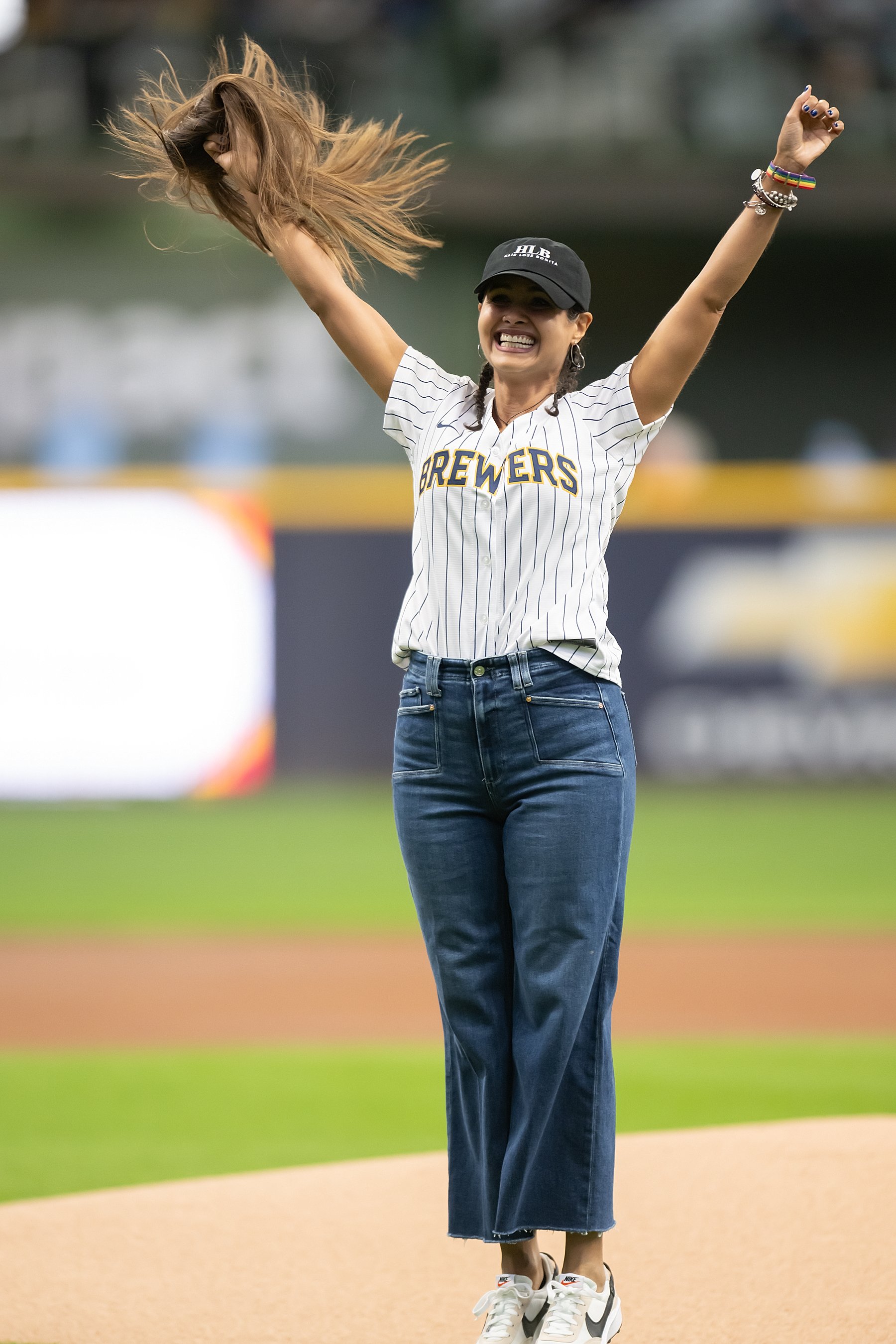 Woman celebrating on a baseball field, wearing a Milwaukee Brewers jersey, jeans, sneakers, baby hair braids and a black cap, with her wig flying and arms raised in joy.