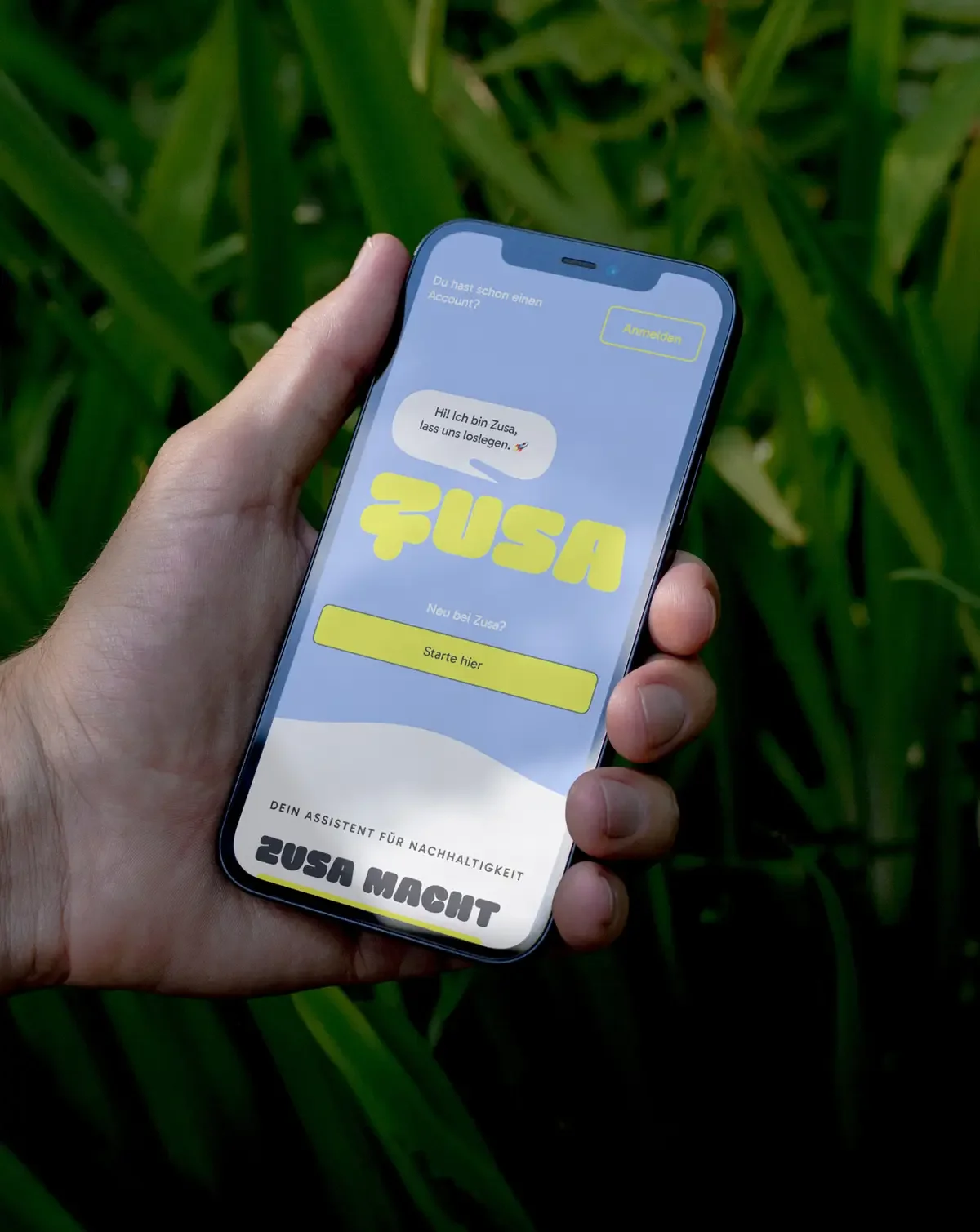 Ein Smartphone in einer Hand zeigt die Startseite der ZUSA-App mit den Worten 'Hi! Ich bin Zusa, lass uns loslegen' und einem Button 'Starte hier' gegen einen Hintergrund aus grünen Pflanzen.