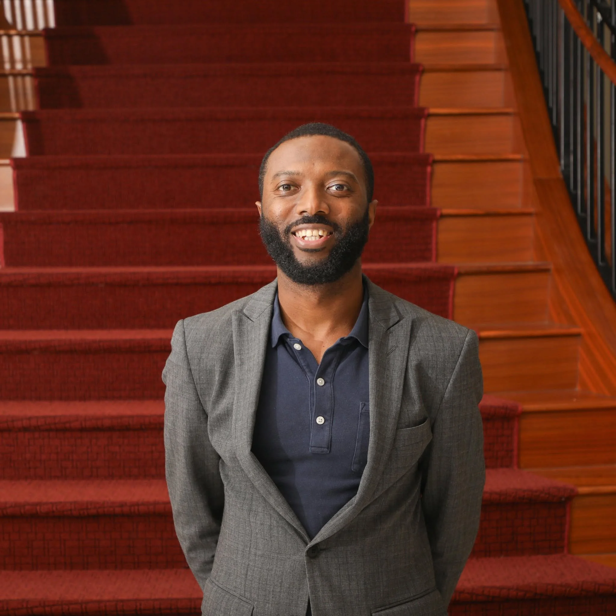 Man smiling in front of carpeted steps, wearing a dark suit