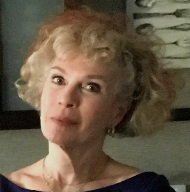 An older woman with curly blonde hair, wearing a blue top, looking directly at the camera.