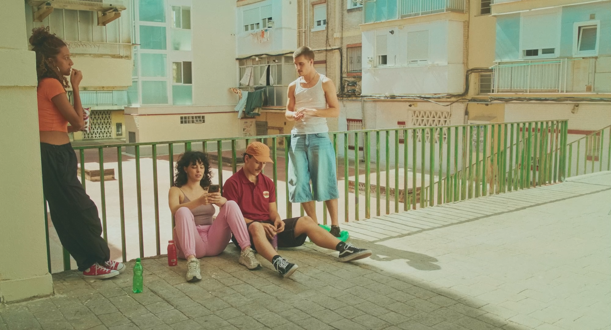 Cuatro personas conversando y relajándose en una terraza al aire libre, dos sentadas en el suelo y dos de pie. Edificios de apartamentos al fondo.