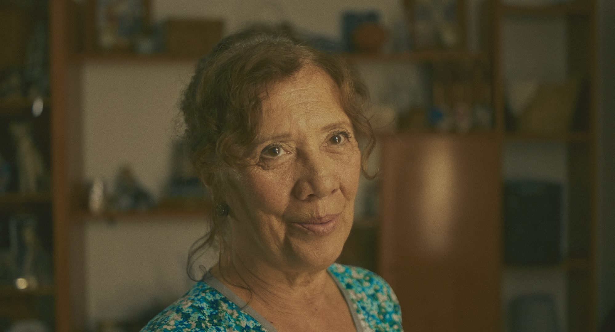 Retrato de mujer mayor sonriendo, con cabello rizado y canoso, en un ambiente interior con fondo difuso. Lleva una blusa azul con estampado floral.