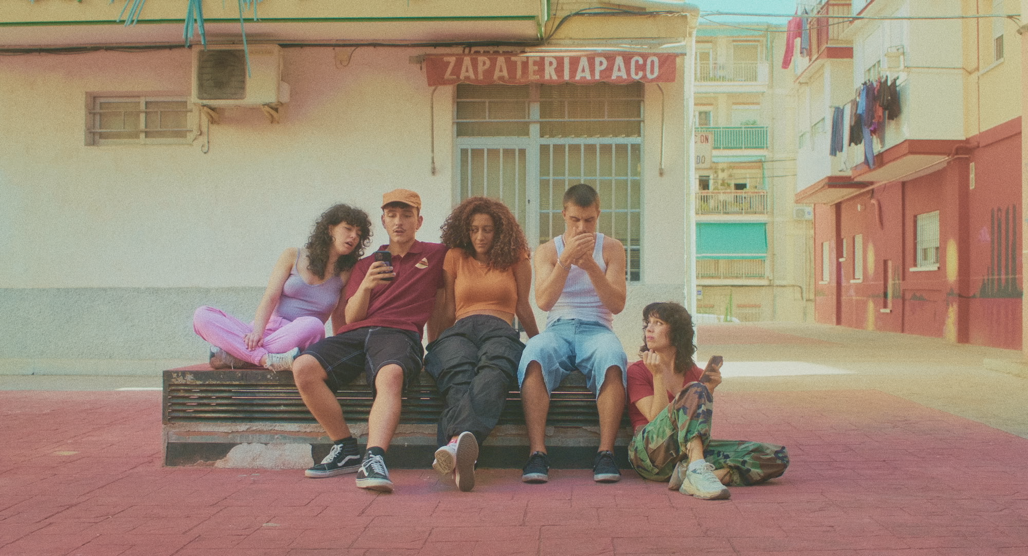 Grupo de cinco jóvenes sentados en una estructura al aire libre frente a una zapatería, cada uno con vestimenta casual, dos están usando sus teléfonos.