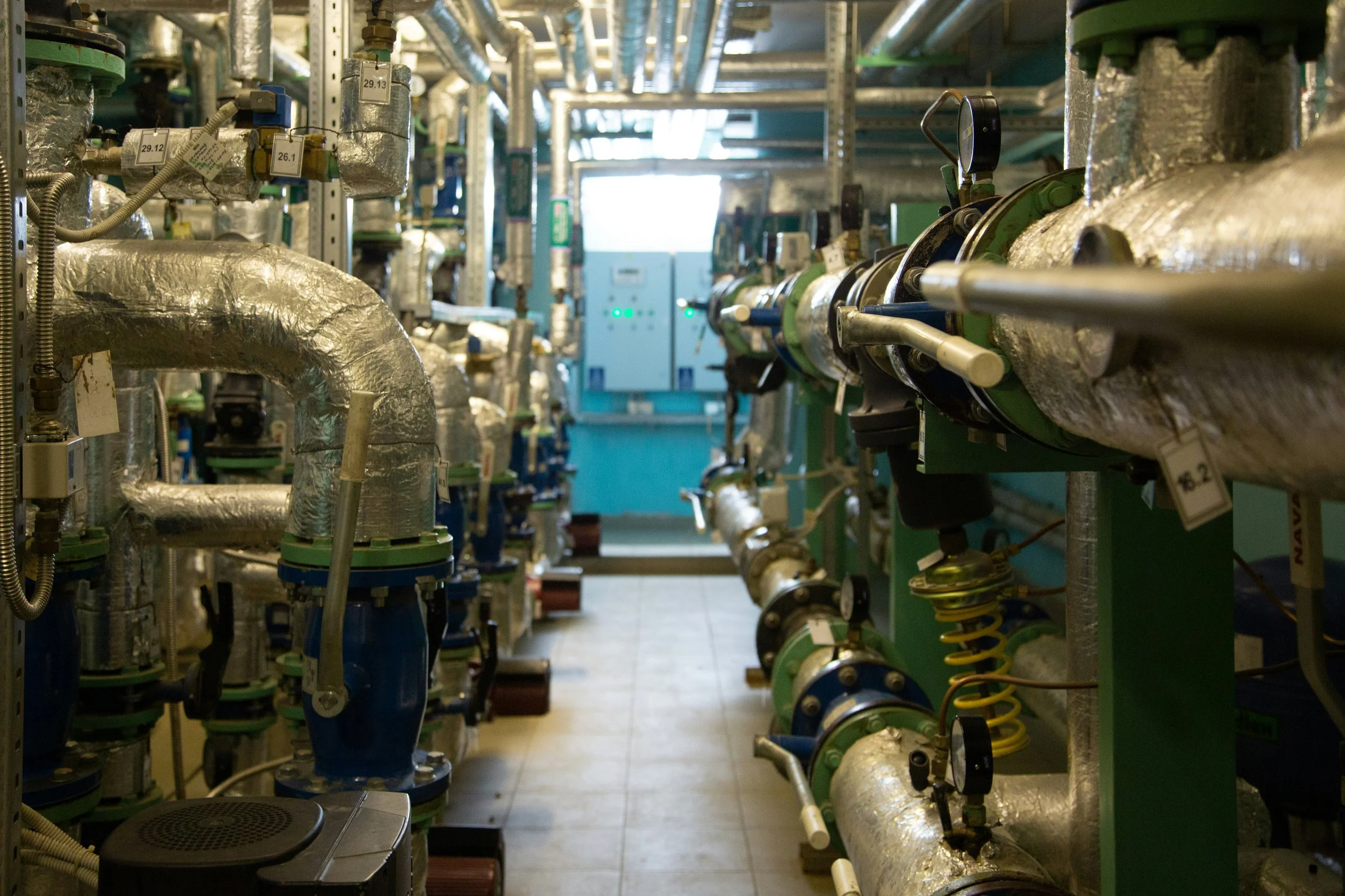 HVAC plant room in a commercial building with insulated pipes, circulation pumps, and a building services control panel