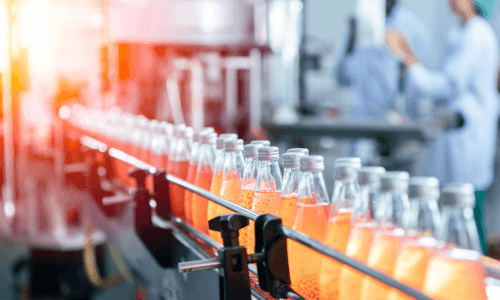 Glass bottles filled with orange-colored liquid moving along a conveyor belt in a factory or bottling plant.