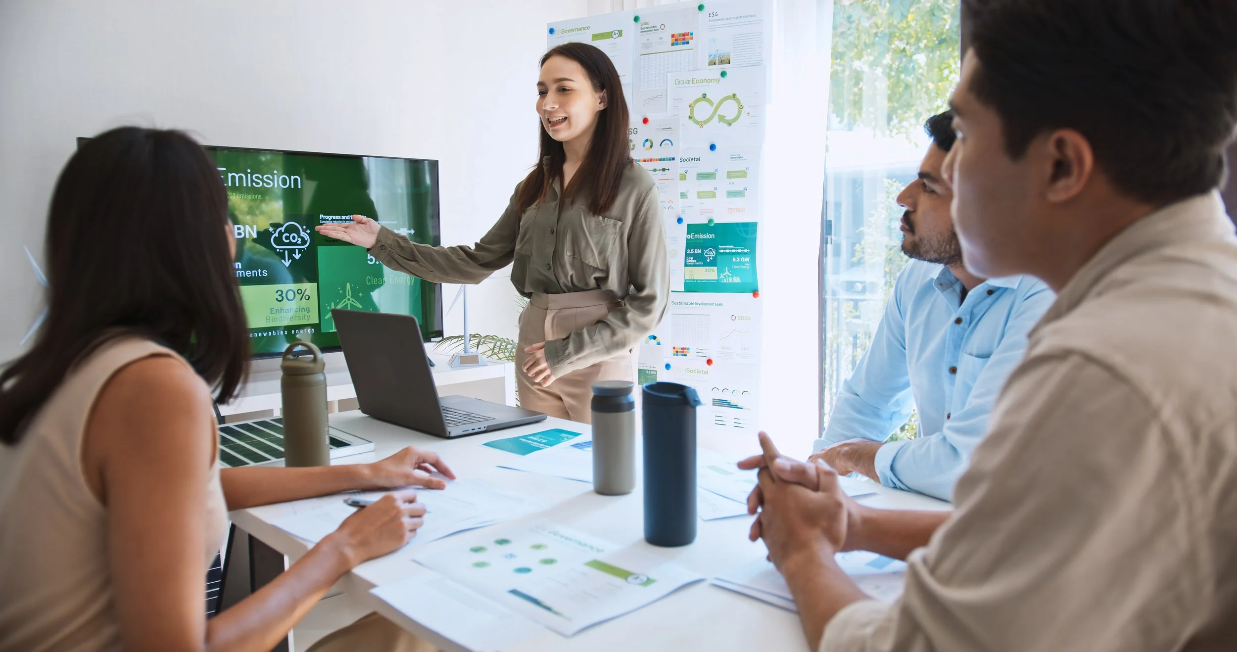 Energy auditors presenting a slideshow on environmental sustainability to three colleagues in a meeting room, with charts and posters about green energy and circular economy visible on the wall and screen.