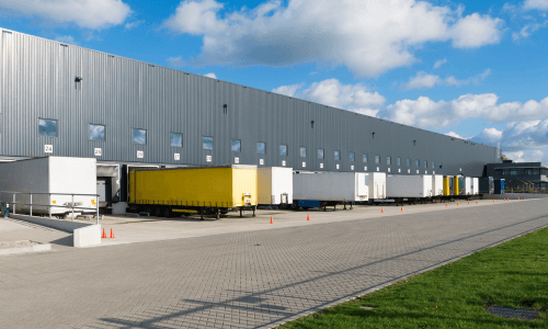 Loading dock with multiple semi-trailers parked outside a large warehouse under a partly cloudy sky.