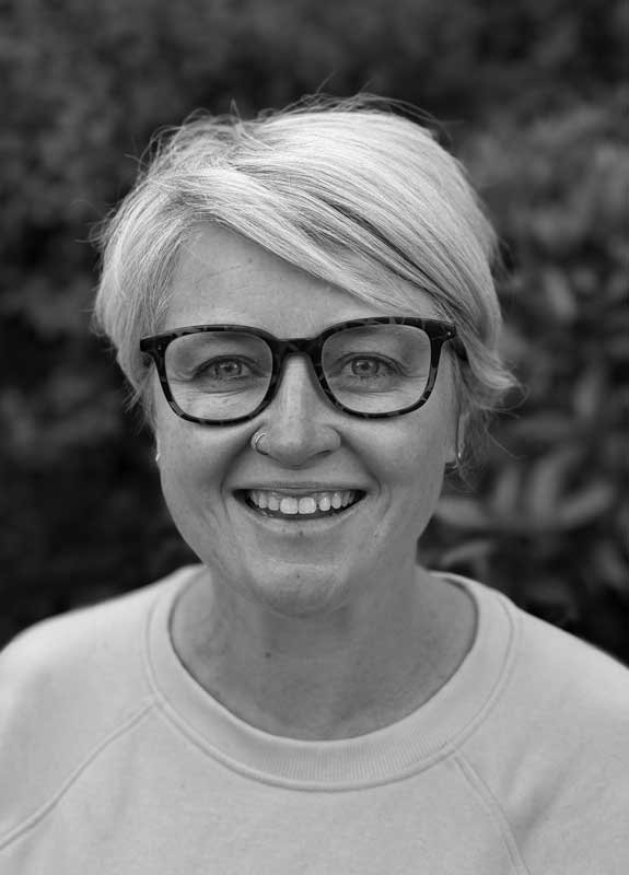 Black and white photo of a smiling woman with short, light-colored hair, wearing large glasses and a nose ring.