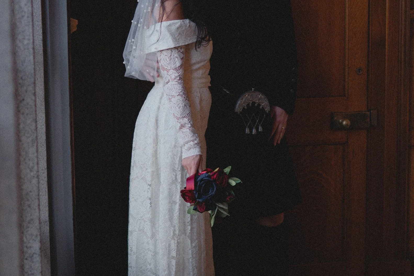 A bride in a white lace wedding dress holding a bouquet of red and black roses.