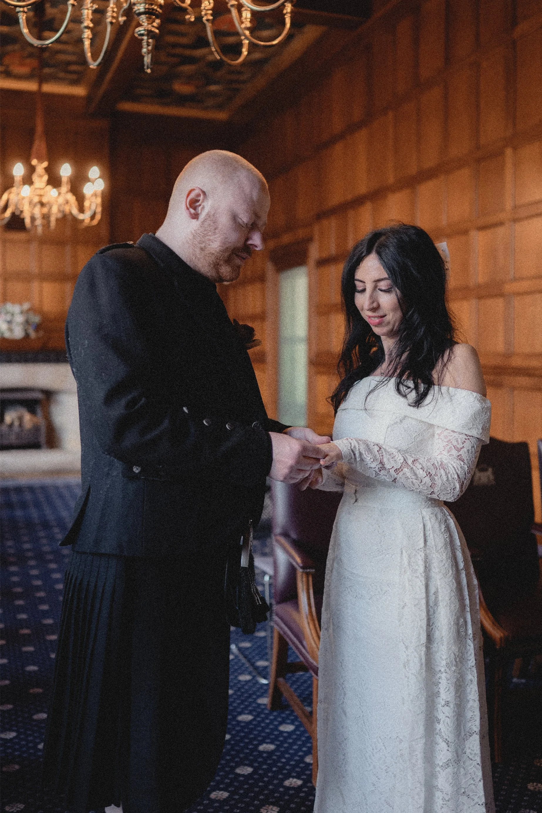 A man and woman are standing indoors with wooden walls. The man is in a black suit and the woman is wearing a white lace wedding dress. They are holding hands and looking down, possibly during a wedding ceremony.