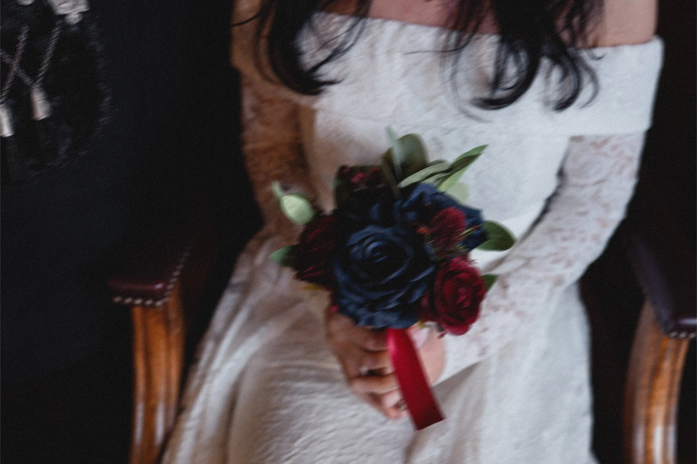 A woman in a white lace dress holding a bouquet of red and dark blue roses with green leaves, sitting on a wooden chair with dark upholstery.