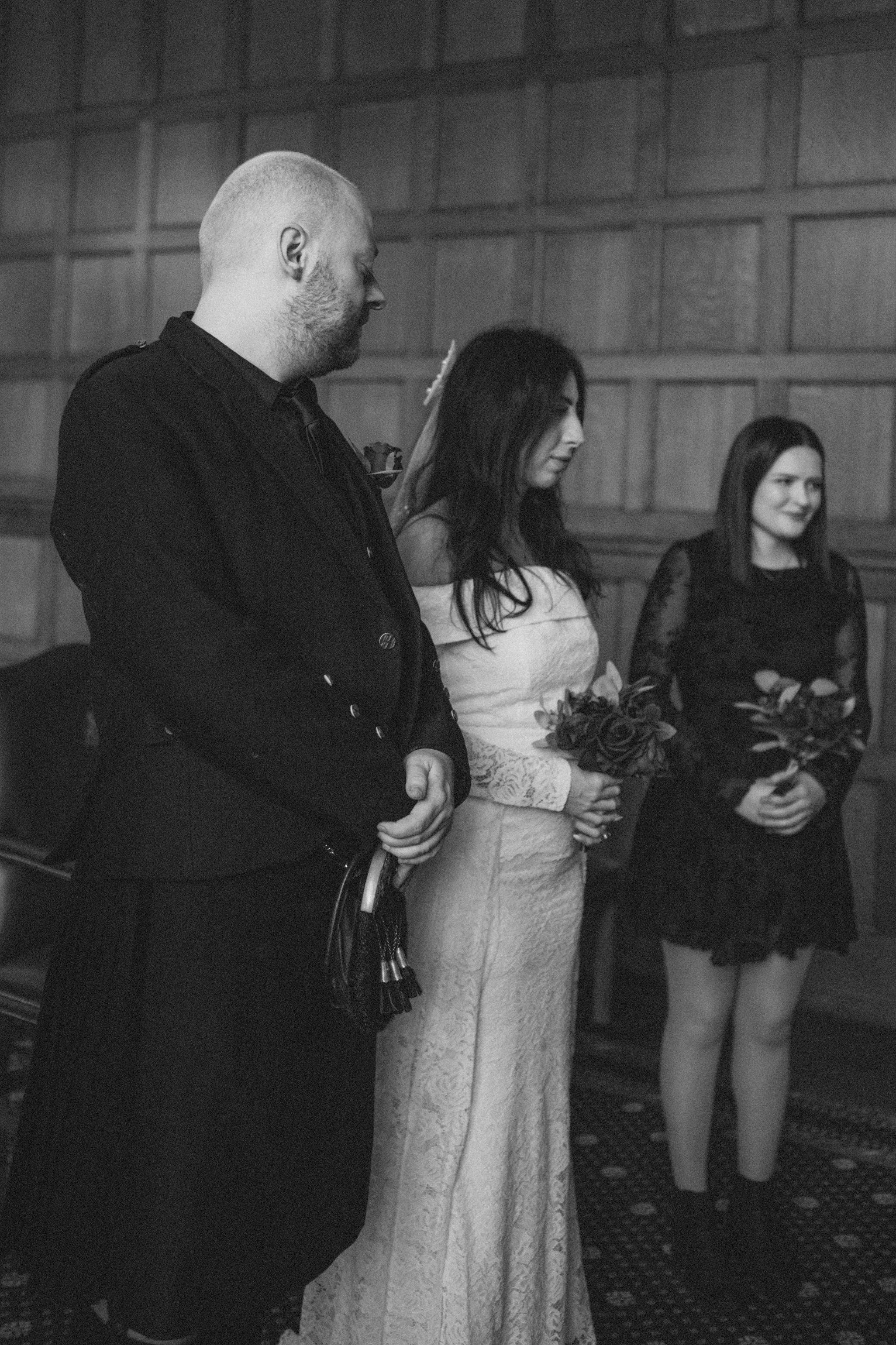 Black and white photo of a wedding ceremony showing a man, a woman in a wedding dress, and a woman in a black dress standing side by side, holding bouquets of roses, inside a wood-paneled room.