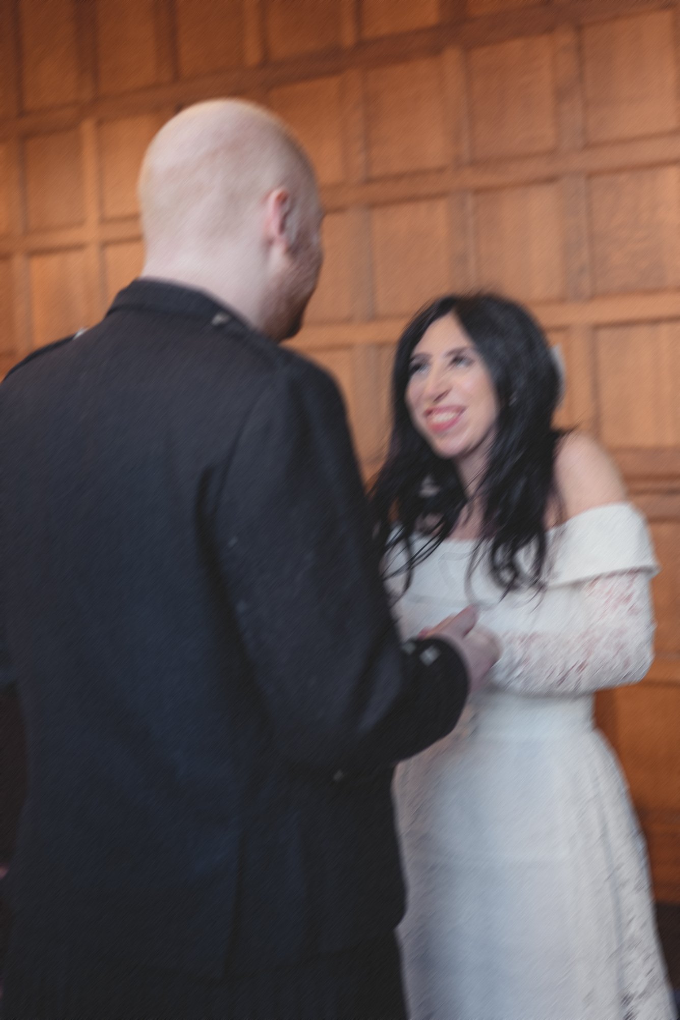 A man with a bald head in a black suit and a woman with long dark hair in a white dress standing close together, smiling, indoors with wooden wall panels in the background.