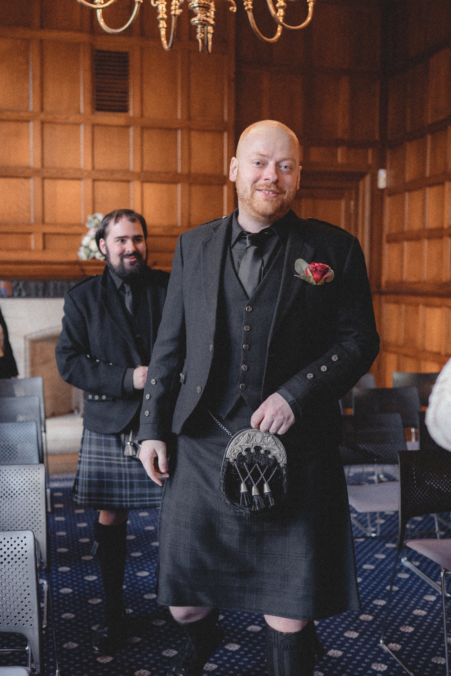 Two men in formal Scottish attire, including kilts, standing in a wood-paneled room with a chandelier. The man in the foreground is smiling, holding a sporran, and has a boutonniere on his jacket. The man in the background is also smiling, wearing a 