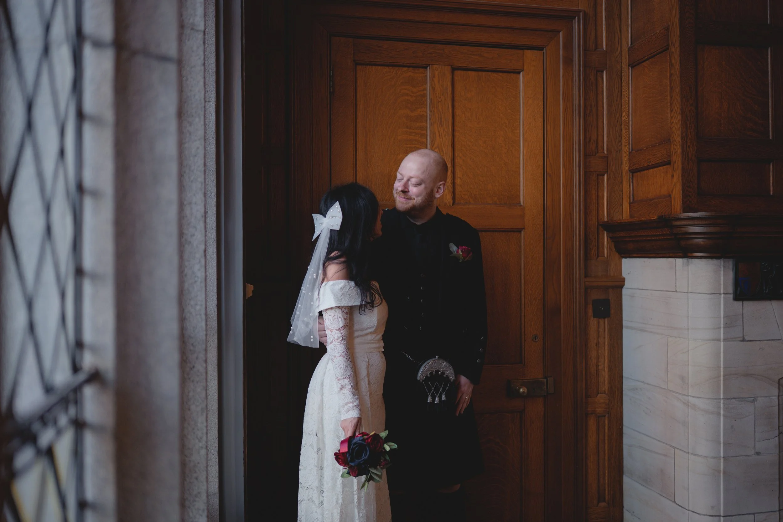 A bride and groom standing close inside a wood-paneled room, sharing a moment of intimacy. The bride wears a lace wedding dress with a veil and holds a bouquet of red and dark flowers. The groom wears traditional Scottish attire with a black jacket a