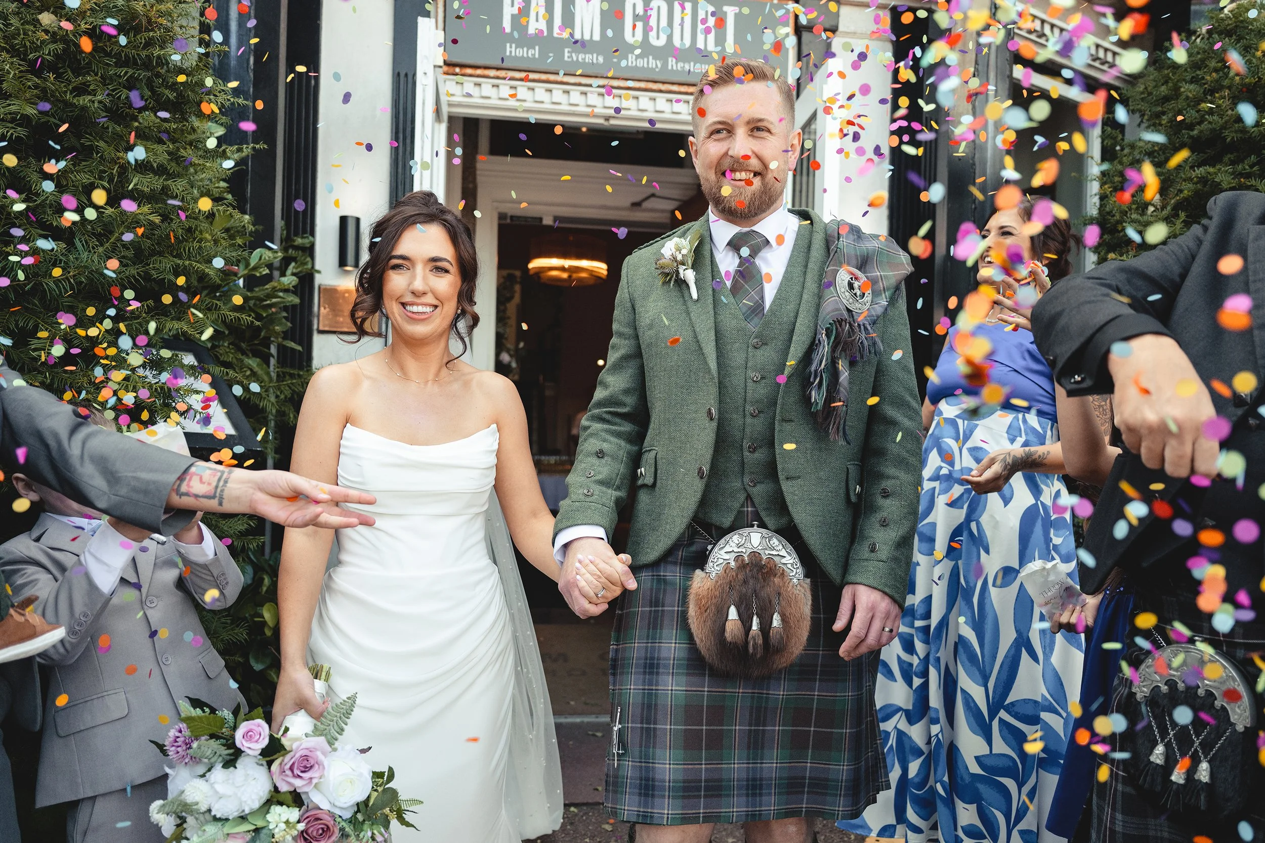 A bride and groom holding hands and smiling at their wedding celebration, surrounded by colorful confetti, with a wedding party and guests in the background outside a venue with greenery and a sign for a hotel and events.