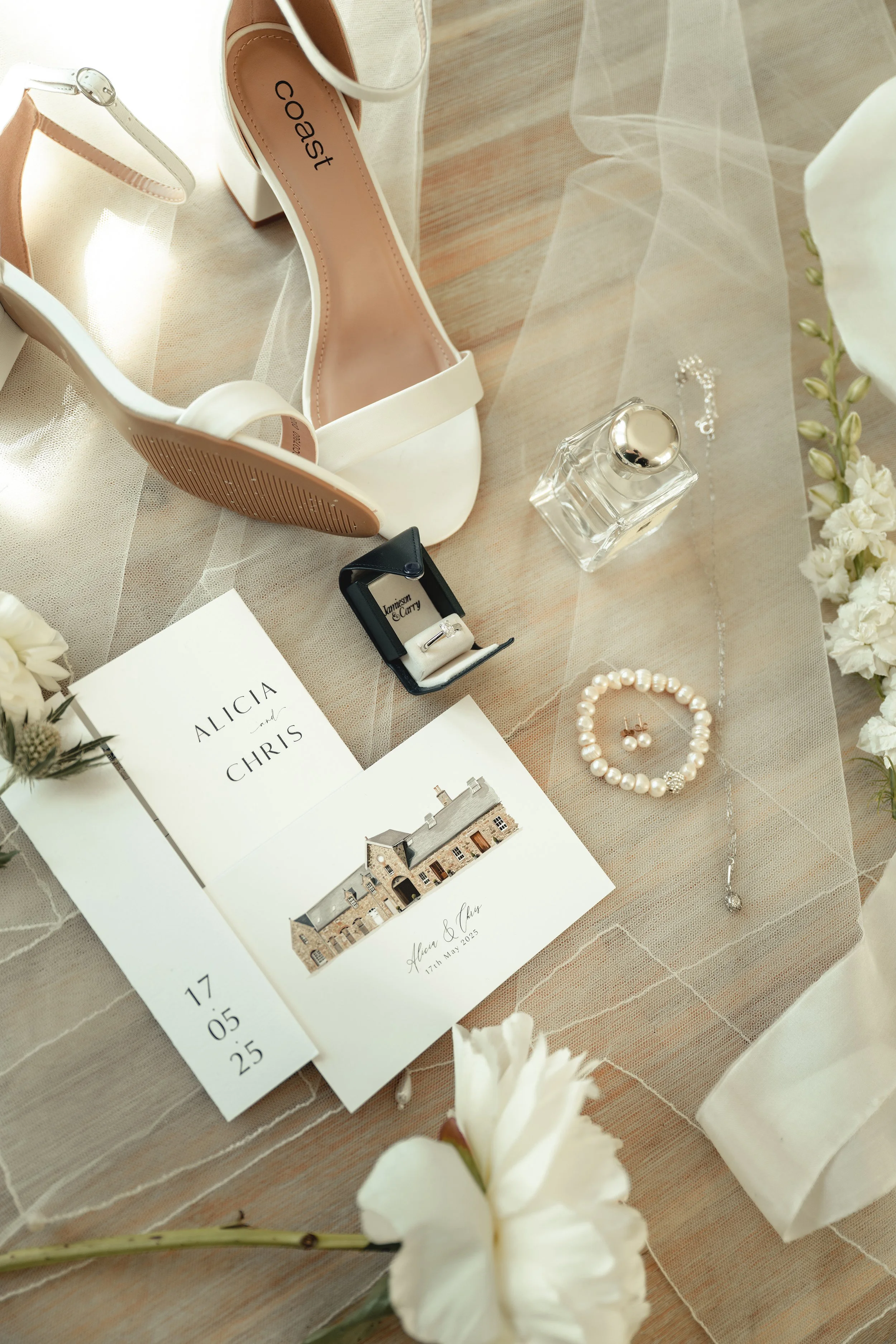 A pair of white high-heeled shoes, a ring in a black box, a bottle of perfume, a pearl necklace and earrings, a wedding invitation, and a boarding pass for Alicia and Chris's wedding on May 17, 2023, are arranged on a beige fabric surface, surrounded
