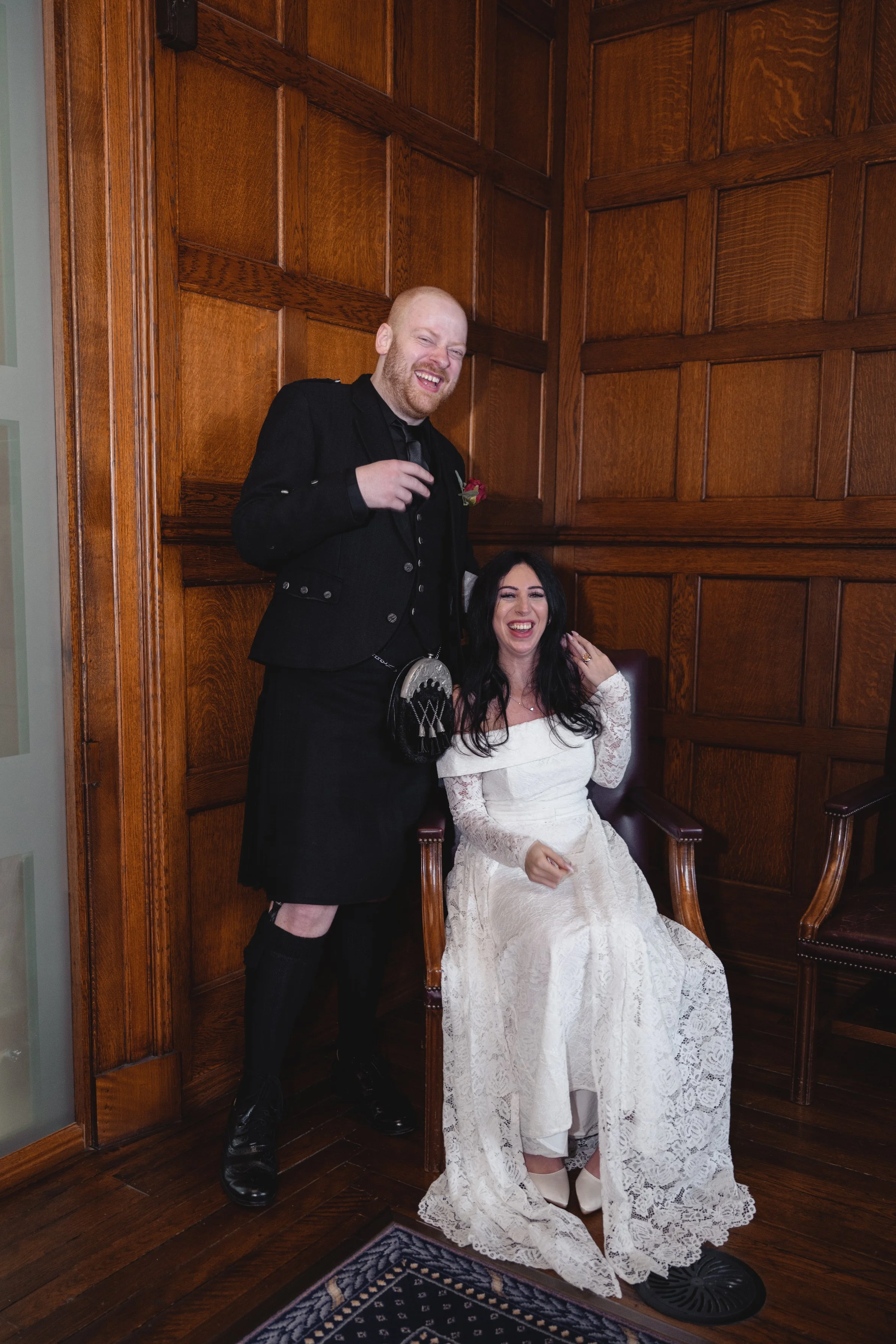 A man in traditional Scottish attire, including a kilt and black jacket, stands beside a seated woman in a white lace wedding dress, both smiling and laughing in a wood-paneled room.