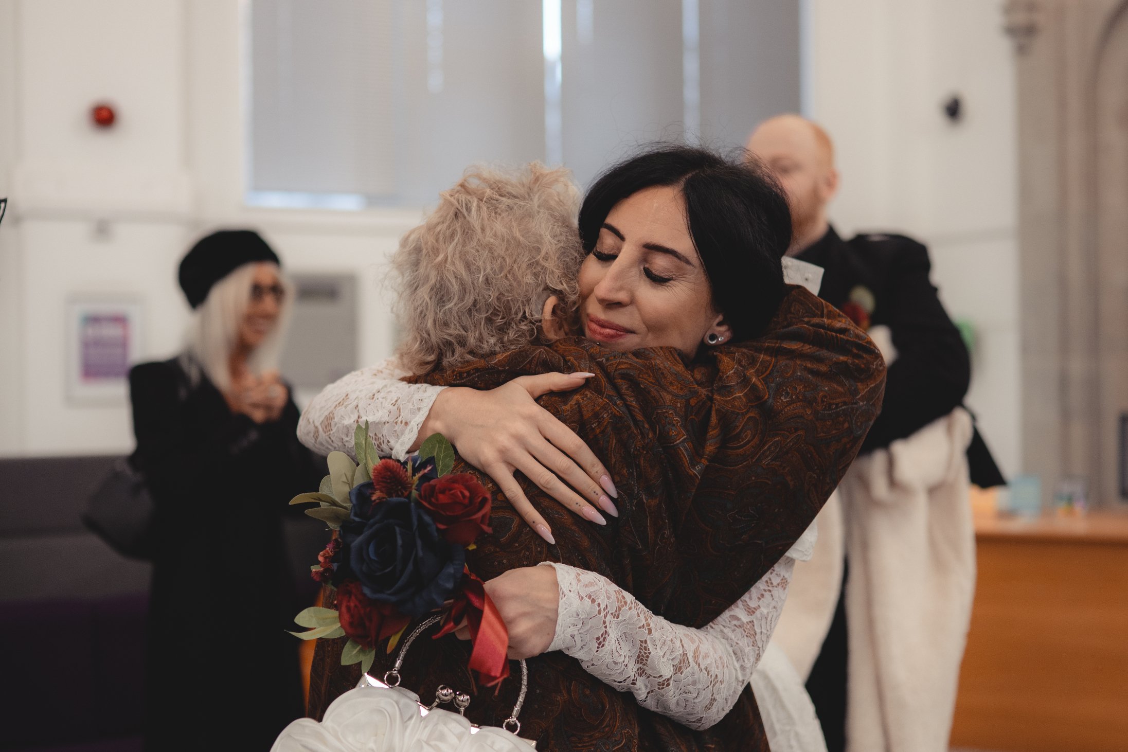 Two women embrace warmly in a hug, with one woman holding a bouquet of red, blue, and purple flowers. Several other people are in the background, standing and chatting in a well-lit indoor setting.