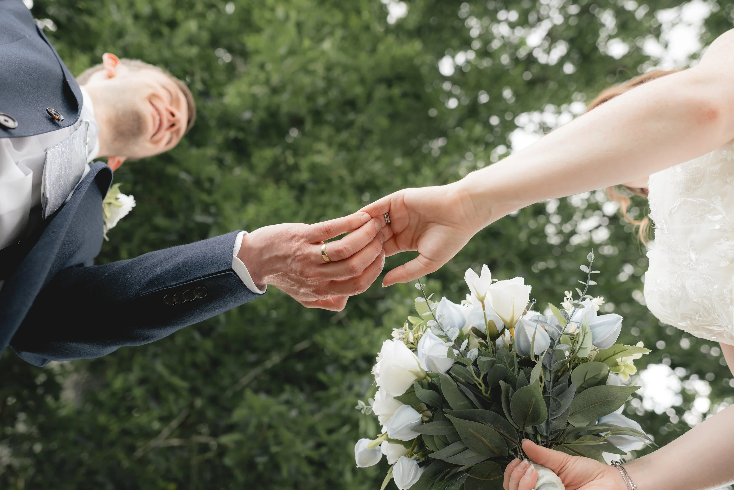 A bride and groom holding hands near a bouquet of white and green flowers, with hand jewelry visible, in an outdoor setting with trees in the background.
