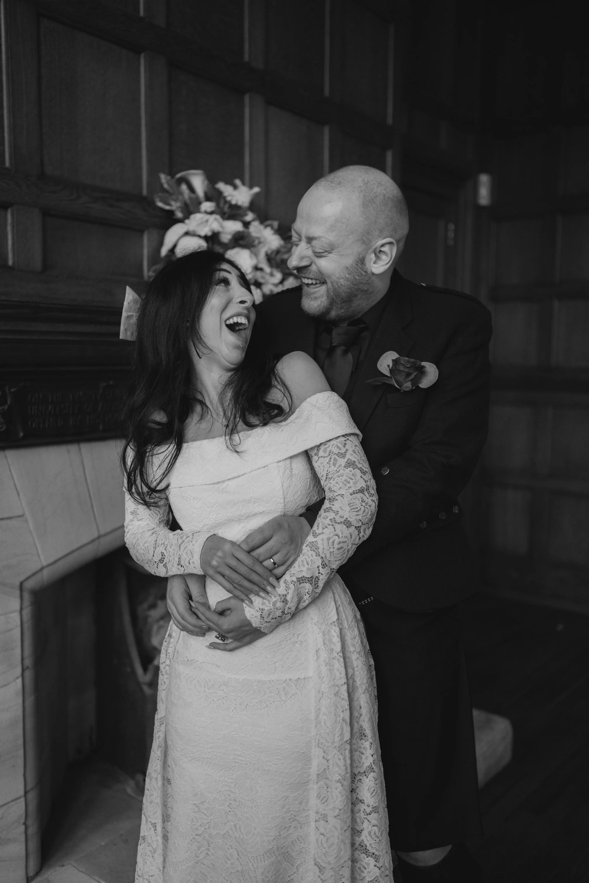 A bride and groom laughing and embracing during their wedding ceremony in a wood-paneled room, with flowers in the background.