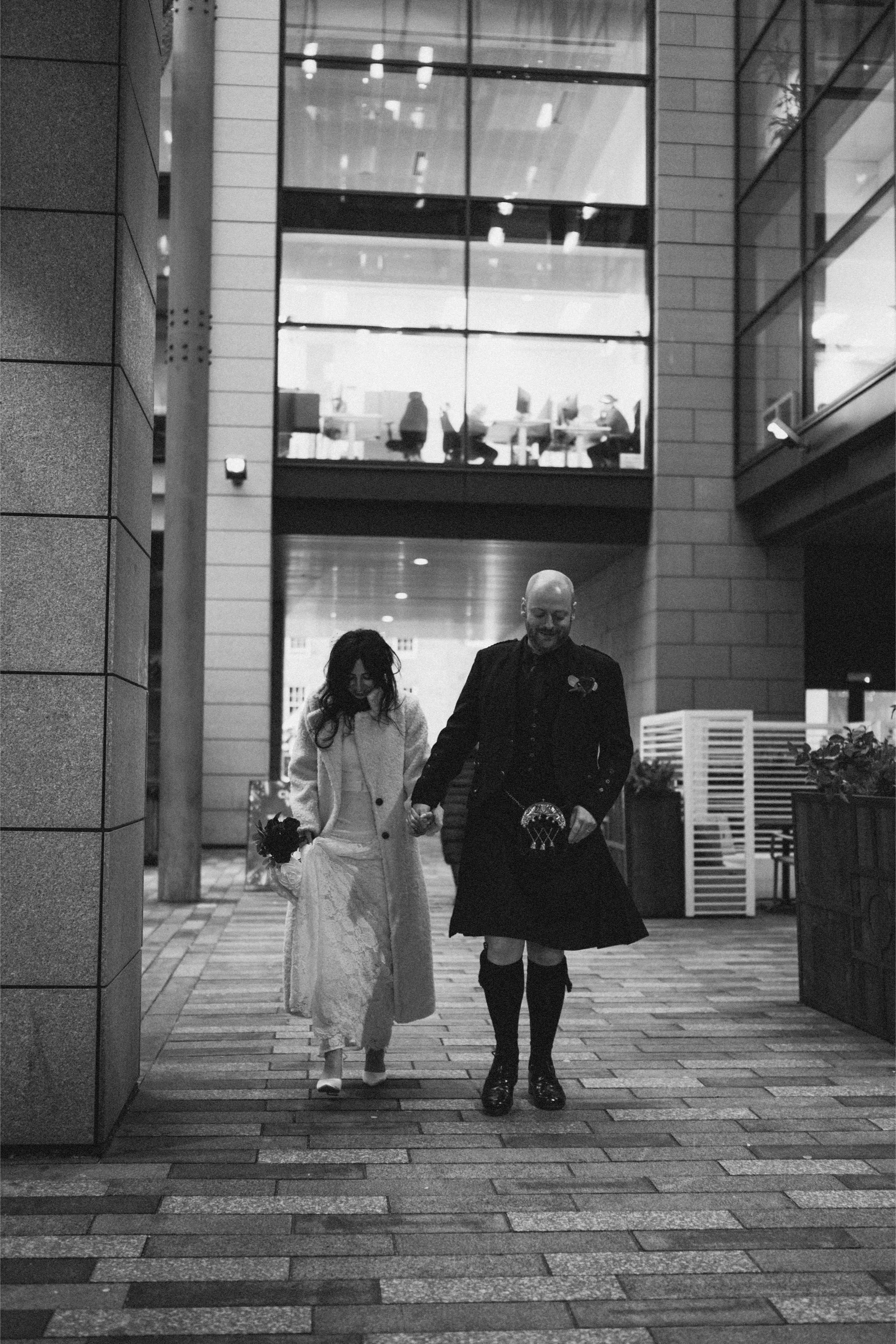 A couple, a woman in a white dress and a man in a kilt, walking hand in hand through an urban area at night, with office buildings in the background and people working inside.